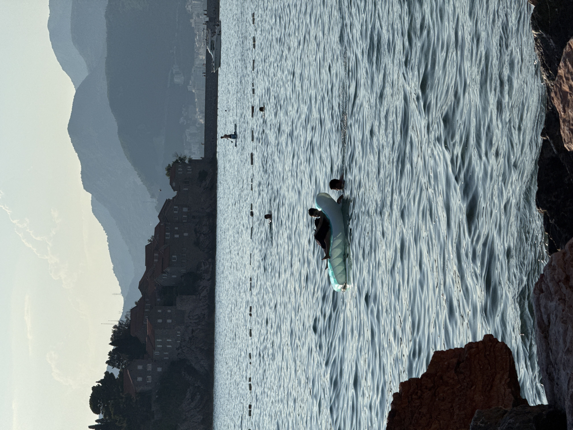 This media file captures a serene scene on the water near Sveti Stefan, Montenegro. The image is oriented vertically, with the water filling the majority of the frame and creating a textured surface of ripples.
In the foreground, a person is seen on a paddleboard. They are seated and appear to be paddling, enjoying the calm waters. Further out on the water, there are several buoys or markers spaced at intervals, suggesting a designated swimming or water sports area. In the distance, a few other individuals can be seen, one standing on what might be another paddleboard or a small boat.
The background reveals a coastal landscape with a mountainous silhouette fading into the distance. Nestled on the rocky coastline is a cluster of traditional stone buildings with red-tiled roofs, characteristic of the area around Sveti Stefan. The sky is hazy and light, suggesting either early morning or late afternoon light. The overall mood is peaceful and tranquil, with the gentle movement of the water and the distant, picturesque scenery contributing to a sense of calm. There is no visible text in the image.