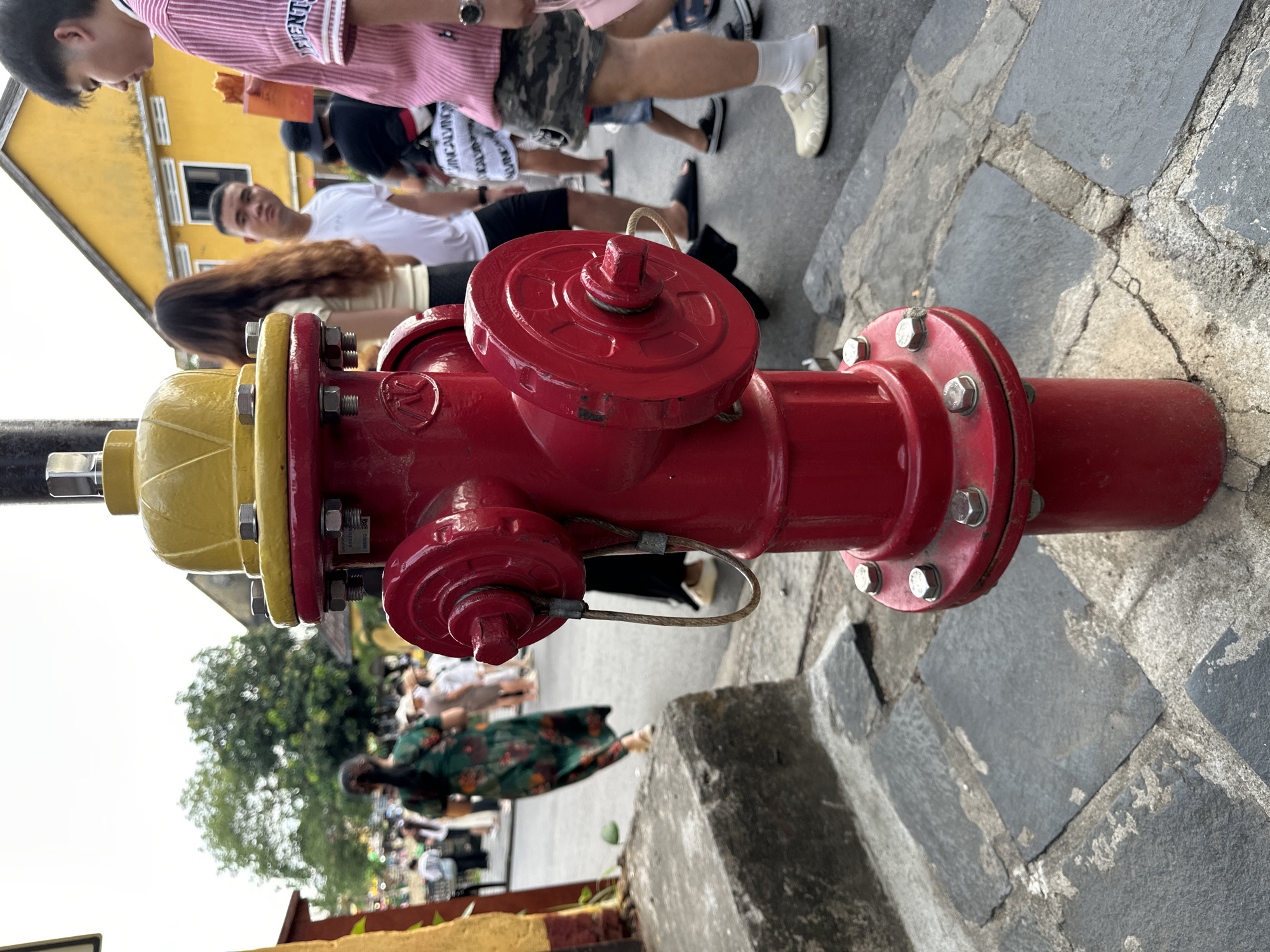 A close-up, low-angle shot features a red fire hydrant with a yellow top positioned on a cobblestone street in Hội An Ward, Vietnam. The hydrant has two side nozzles and a large cap on top, secured with bolts. In the background, several people are walking in a public area. To the left, a woman in a green floral dress walks away from the camera. In the center, a man in a white t-shirt looks towards the right, and a woman with long brown hair stands beside him. To the right, a person in a pink and white striped shirt is visible, with a young person in a white t-shirt and camouflage shorts walking past. The environment appears to be a street scene with buildings and trees.