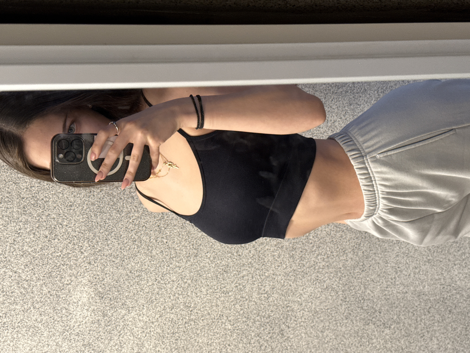 A young woman is captured in a mirror selfie, holding a smartphone to take her picture. Her reflection, oriented vertically in the mirror, shows her from approximately the chest down to just above the knees. She has dark hair, with parts visible around her face, and blue or grey eyes looking directly into the phone's camera.
She is wearing a simple black ribbed crop top or camisole, revealing her midriff, and light grey sweatpants with an elasticated waist. Accessories include a gold-colored necklace with a small charm, a dark beaded bracelet on her right wrist, and a silver-toned ring on her left hand's ring finger. Her fingernails are manicured in a light, neutral shade.
The smartphone she is holding is dark-colored, possibly black or dark grey, and features a prominent white circular ring on its case, suggestive of a MagSafe-compatible design. The phone's multi-lens camera module is also visible.
The setting appears to be indoors, likely a room. The floor visible in the reflection is covered with a light grey, speckled carpet or rug. A horizontal white structure, possibly a window sill, wall trim, or part of a mirror frame, runs across the top portion of the image. The lighting is even, suggesting an indoor environment without strong natural light indicators. There is no visible text in the image. The activity is clearly a self-portrait taken in a mirror. While the context states Sofia, Bulgaria, there are no specific visual cues within the image to confirm this location.