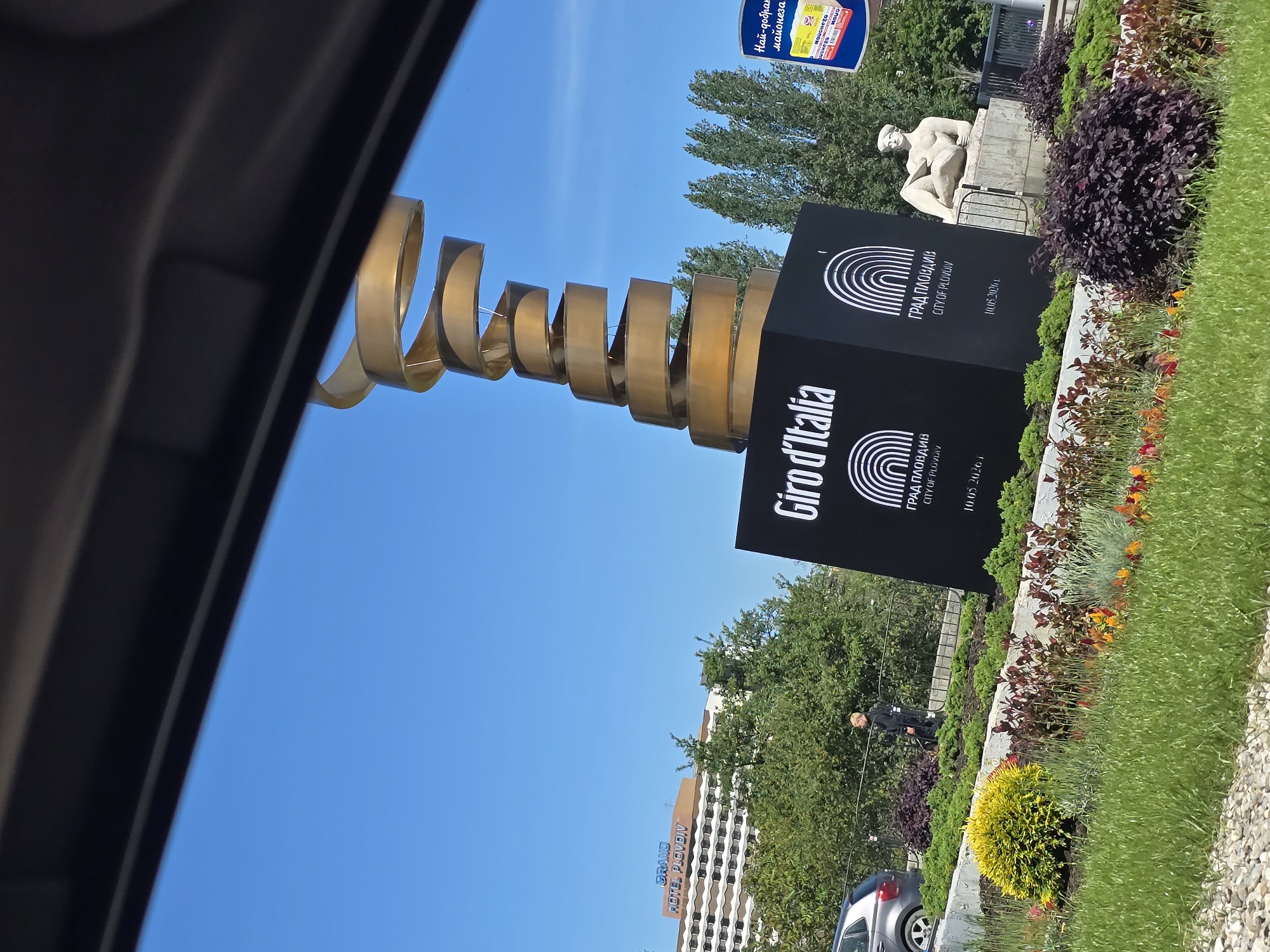 Here is a description of the image:
The image depicts a street scene in the city of Plovdiv, Bulgaria. The main subject is a monument commemorating the Giro d'Italia. The monument features a black cubic base with the logo and inscriptions related to the city of Plovdiv. Atop the cube, a metallic, spiraling structure rises, resembling a ribbon or road.
In the background, there is a statue of a seated nude male figure on a stone plinth, behind the monument. In the foreground, landscaping elements are visible including grass, flowers, and other greenery, separated by a low stone wall. A person wearing dark clothing is discernible among the greenery.
The buildings in the background include a tall hotel identified as "Grand Hotel Plovdiv".
The composition of the image suggests it was taken from inside a vehicle, with parts of the car frame visible. The sky is clear and blue. A billboard can also be seen above and behind the monument.