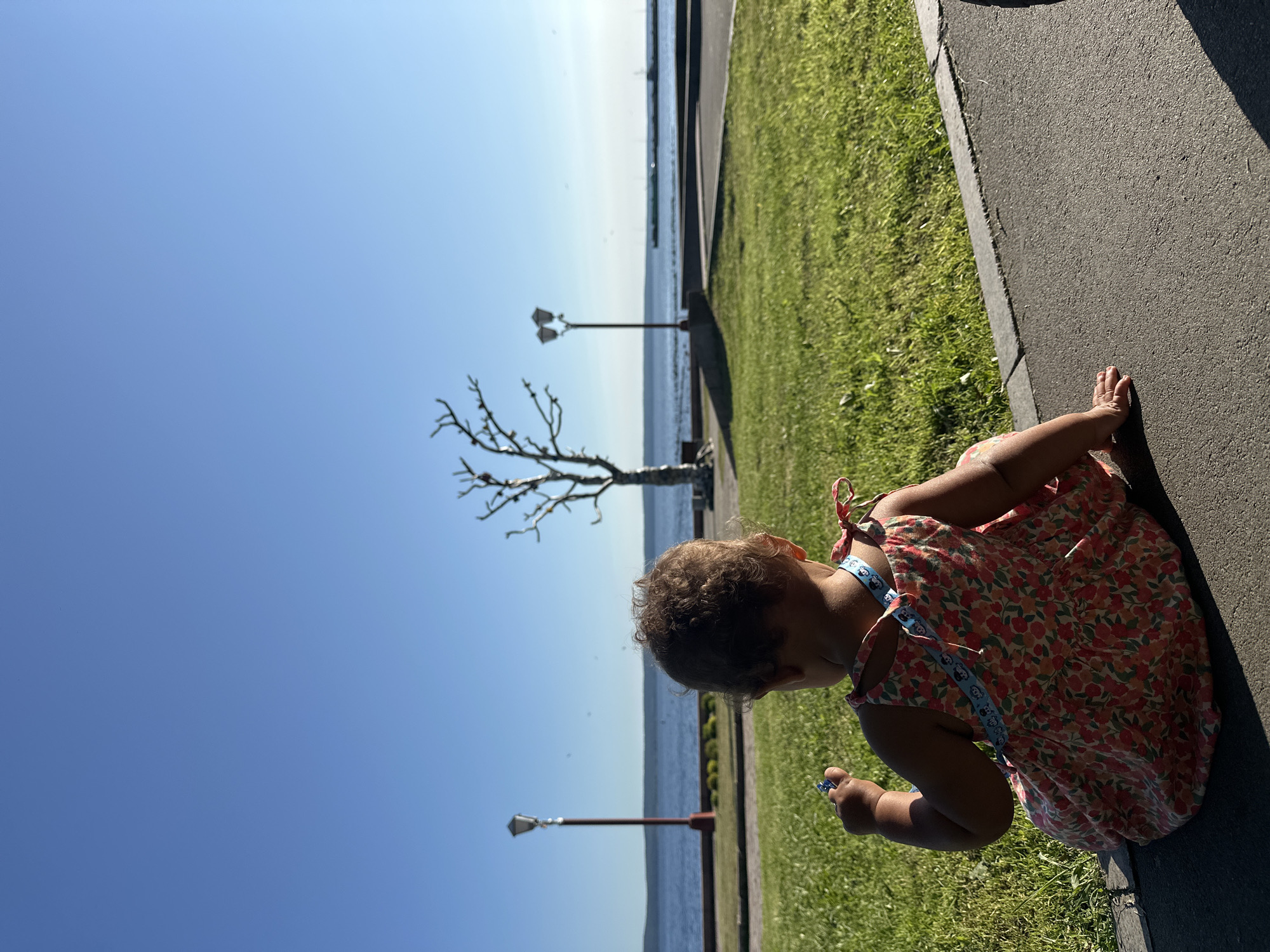 A young child sits facing away from the camera on a paved surface, looking towards a body of water, likely Lake Onega, with a clear blue sky above. The child is wearing a floral-patterned dress and has curly brown hair. In the background, there is a grassy area, a decorative tree sculpture, and a lamppost. Further in the distance, the calm water stretches to the horizon, with faint structures visible on the opposite shore. The scene depicts a peaceful outdoor setting, possibly a park or waterfront promenade in Petrozavodsk, Russia.