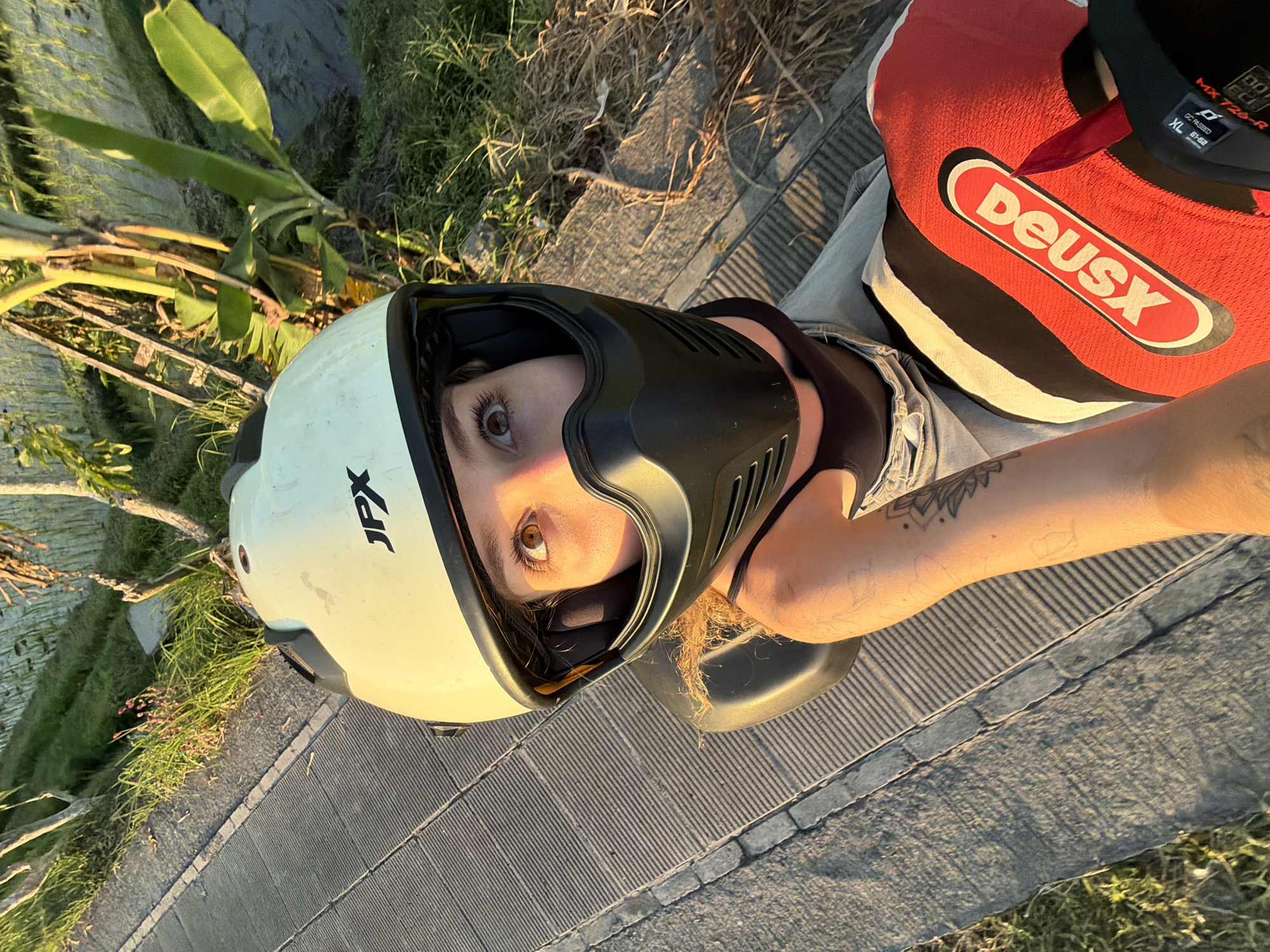 A POV shot captures a young woman wearing a cream-colored motorcycle helmet with a black face mask and goggles, looking upwards towards the camera. Her eyes are open, and she appears to be smiling slightly. She is seated on a motorcycle, with her right arm extended, holding a red and white racing glove with the "Deus Ex Machina" logo visible. Her left arm, with a tattoo on her forearm, rests on the motorcycle's seat. She is wearing a black tank top and light-colored pants.
The background suggests a rural or natural setting, possibly a rice paddy field, with lush green vegetation and banana trees visible to the right. To the left, a textured path or walkway made of concrete slabs runs parallel to a grassy area. The scene is illuminated by the warm glow of what appears to be sunset or sunrise. The location is identified as North Kuta, Indonesia.