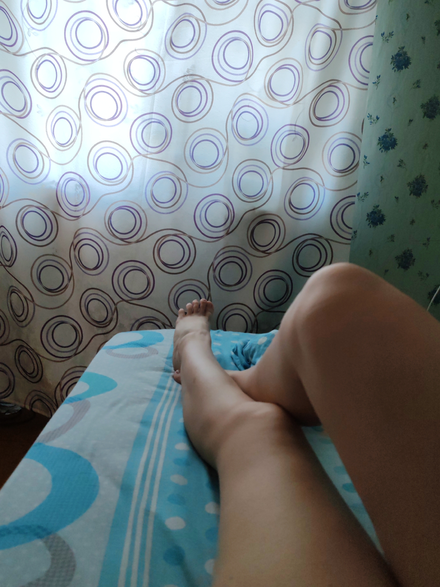 The image displays a close-up, first-person perspective of a person's lower legs and feet resting on a bed in an indoor domestic setting. The visible parts of the legs and feet have light skin tone. The person is lying on light blue bedding patterned with white and darker blue abstract shapes, including arcs, stripes, and dots.
In the background, two distinct sections of window coverings or wall decor are visible, illuminated by natural light. The main section, to the left and center, features a sheer or translucent curtain with an abstract design of concentric purple and brown circles connected by wavy lines against a white background. To the right, a second curtain or wallpaper is present, characterized by a light greenish-blue base adorned with a repeating pattern of dark blue floral clusters and scattered leaves. The scene is situated in the city of Chukhlym (Чухлэм), Russia.