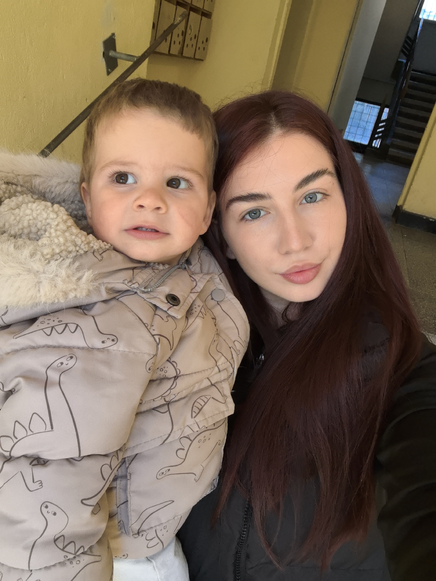The image shows a woman holding a young child. The woman has long, auburn hair and light blue eyes. The child is wearing a beige jacket with a dinosaur pattern. The background shows a hallway with yellow walls and a staircase. Based on visual clues from the wall-mounted mailboxes and staircase visible in the background, the image was taken in an apartment building in an urban setting. The specific location appears to be in Stara Zagora, Bulgaria, inferred from the user request and the overall architectural style.