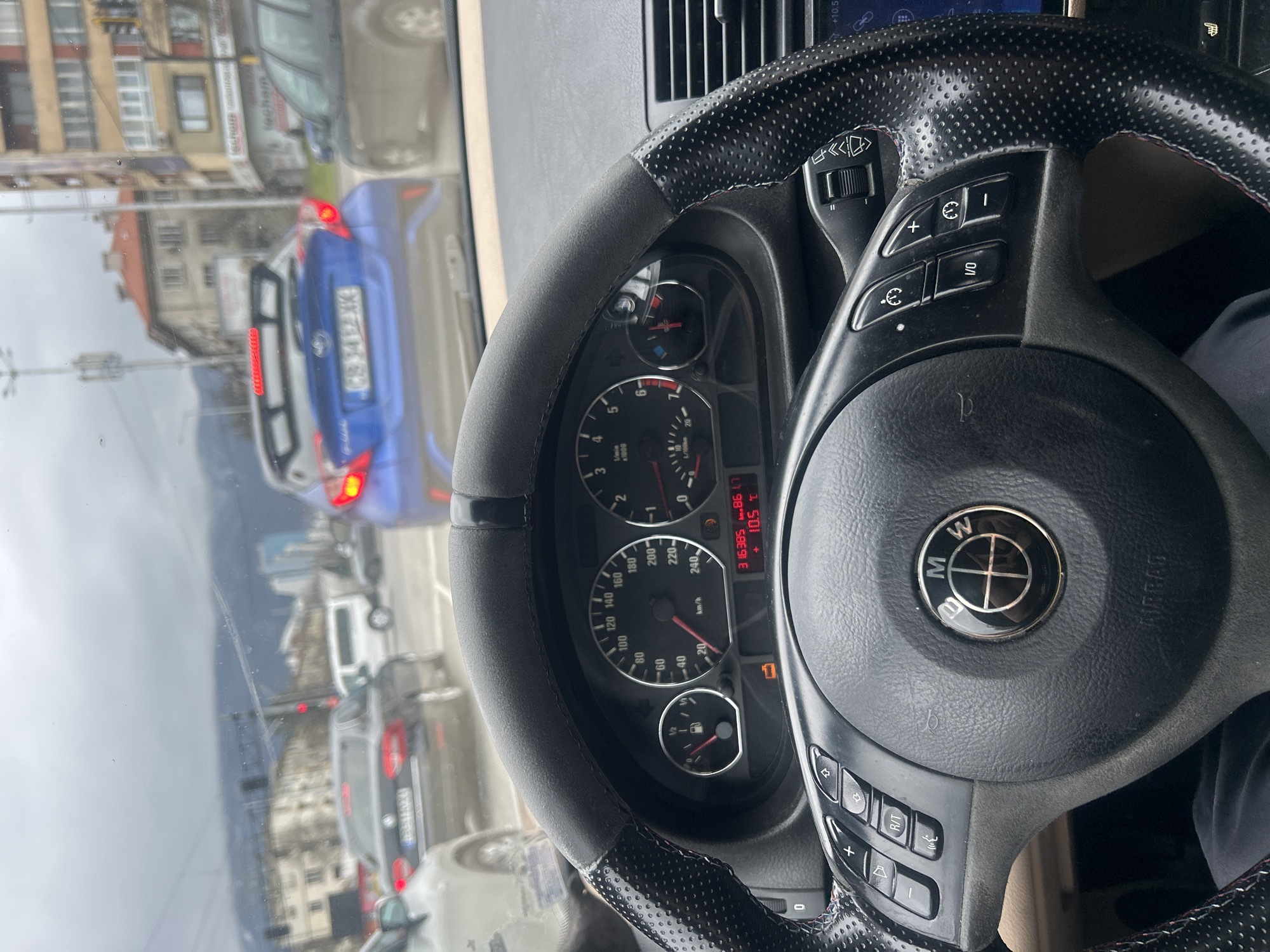 The image depicts the interior of a BMW car, with the steering wheel and dashboard prominently in view. The speedometer indicates a speed of approximately 20 km/h. The mileage is displayed as 316385 km, and the outside temperature is 10.5°C. Through the windshield, a view of a city street is visible. In the reflection, a blue Toyota car is seen with license plate CB 9444 XX. Buildings and other vehicles indicate an urban environment. The presence of signage possibly indicating "lecham" suggests that this scene is located in Sofia, Bulgaria. The image presents a snapshot of driving conditions in Sofia, with traffic and urban scenery.