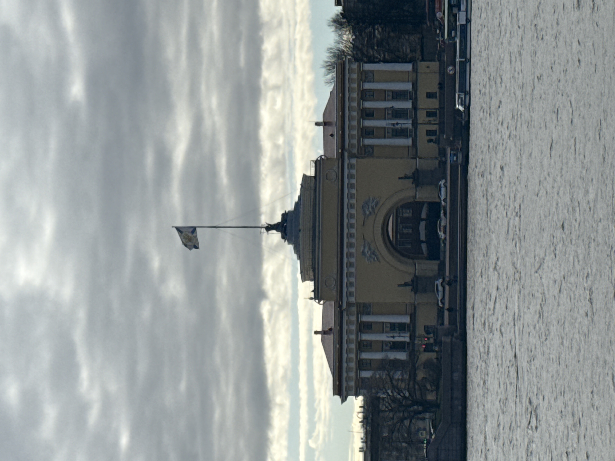 This image captures a waterfront urban scene in Saint Petersburg, Russia.
**Subjects:** The scene is dominated by a grand, light-yellow classical-style building positioned on the left side of the frame. This structure features prominent columns, arched windows, and ornate decorative elements. A flagpole extends from its roof, flying a light-colored flag bearing a dark, indistinct emblem in its center. To the left and right of this central building, other urban structures line what appears to be an embankment or street. A wide body of water, likely a river or canal, fills the right portion of the image; its surface exhibits a rough texture, suggesting either choppy conditions or the presence of ice slush. Several cars are visible parked along the waterfront. The upper part of the image is filled with an overcast sky, displaying various shades of grey clouds. Bare trees, indicating a cold season, are visible near the buildings.
**Activity or event taking place:** The photograph presents a static urban landscape, depicting a tranquil moment in the city's daily life, without any specific human activities or events taking place.
**Notable details:** The diffused daylight suggests an overcast or cloudy day. The bare trees and the textured appearance of the water are consistent with cold weather, possibly winter or early spring conditions. The classical architecture of the buildings, particularly the prominent yellow structure, is a strong architectural characteristic of Saint Petersburg, Russia. The flag is likely a municipal or regional flag. No decipherable text is visible in the image.