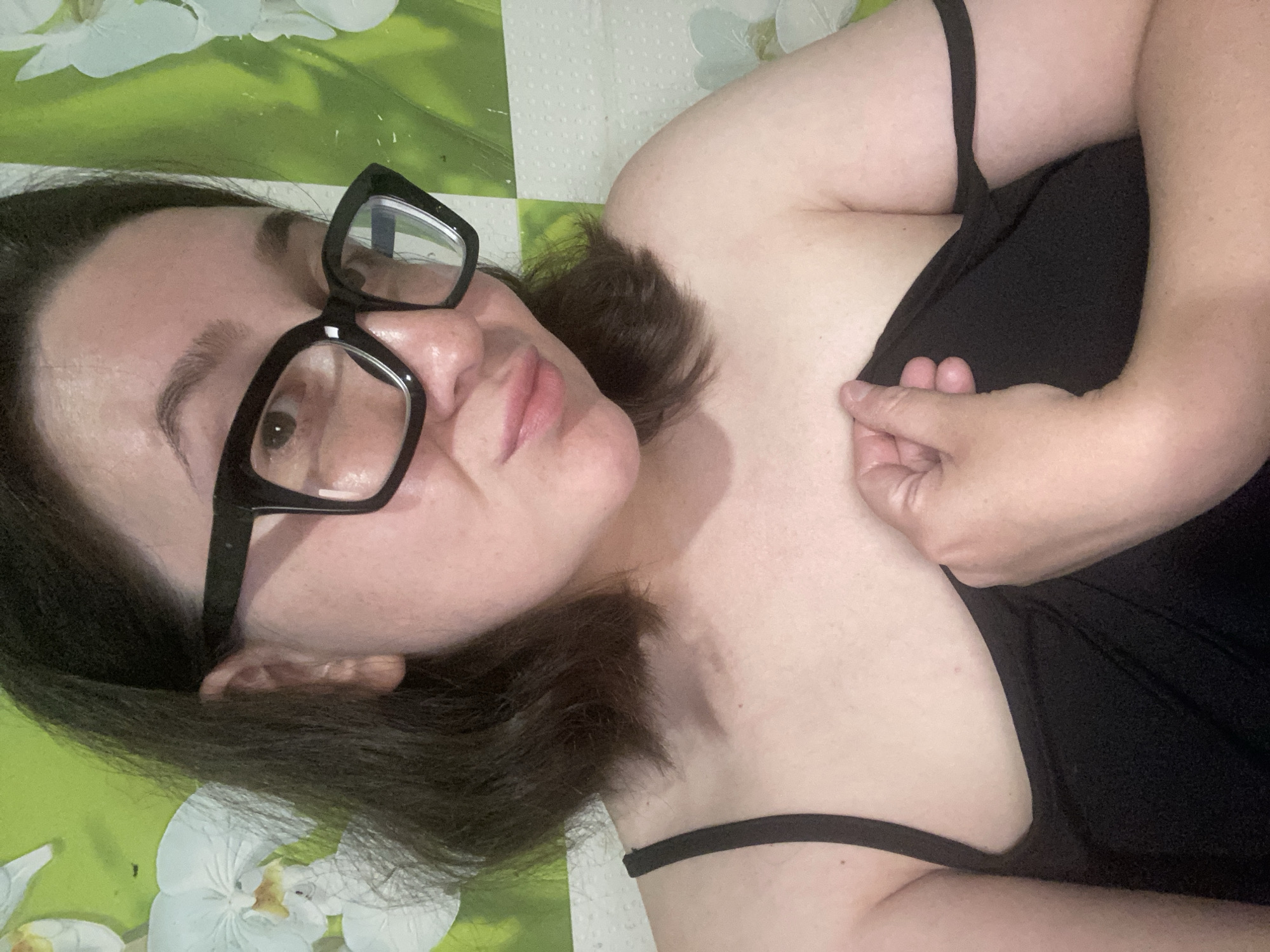 A woman with dark hair and black-framed glasses is lying on a surface with a green and white floral pattern. She is wearing a black top. The location is unknown and cannot be determined from the image.