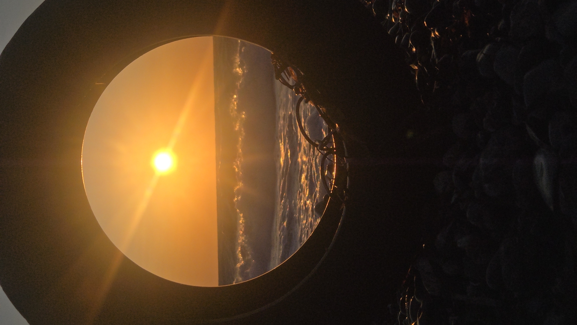 A circular frame, appearing to be the inner edge of a tire, encloses a vibrant sunset over the Aegean Sea. The sun, a bright orb radiating golden light, hangs low in an orange sky, casting a luminous path across the rippling water. Waves with white crests break gently, reflecting the warm hues of the sunset. Within the foreground, dark, rough stones and some coiled rope or debris are visible, partially obscuring the bottom of the circular frame. The overall scene evokes a sense of tranquility and natural beauty, framed by an unexpected, man-made element. The location is the coast of Soroni, Greece.