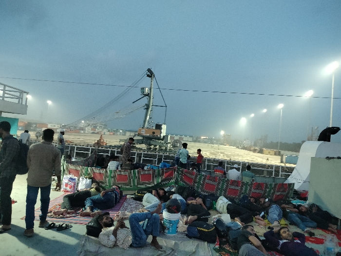 The image shows a group of people sleeping or resting on the deck of a ship at night. The ship is docked at a port facility in Zazira, Bangladesh, which is located at coordinates 23.658 latitude and 90.456 longitude. The sky is overcast and there are several large, brightly lit cranes and other port equipment visible in the distance. The people are lying on blankets, some of them are wearing clothing, some are not. The deck is made of metal and is dirty. The ship's railing is visible in the background. There are no obvious signs of distress or conflict.