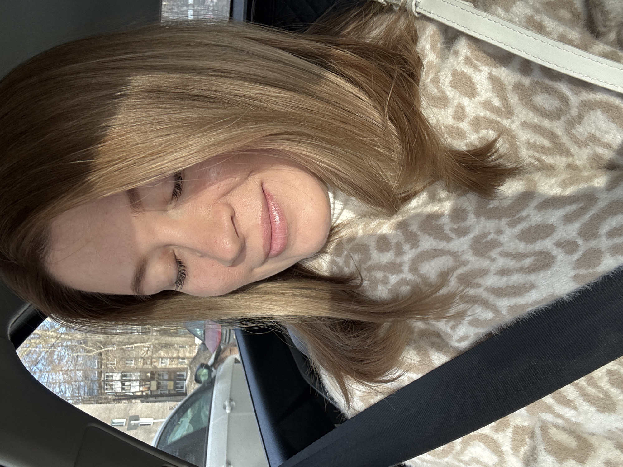 The image depicts a female individual seated as a passenger inside a vehicle in Chaikovsky, Russia. The subject, viewed from the chest up, has light brown, shoulder-length hair and fair skin with visible freckles on her nose and cheeks. Her eyes are closed, and a subtle smile is present on her face. She wears a cream and beige leopard-print turtleneck sweater and is secured by a black seatbelt diagonally across her left shoulder and chest. A white strap, possibly from a bag, is visible on her right shoulder. Direct sunlight illuminates the left side of her face and hair. Through the vehicle's window, an urban environment is visible, featuring multi-story grey residential buildings with multiple windows and balconies, characteristic of the city. Bare deciduous trees are interspersed between the structures. Parked vehicles, including a white car and a partially visible red car, are present in the background. Patches of snow are visible on the ground, and the sky is clear and blue.