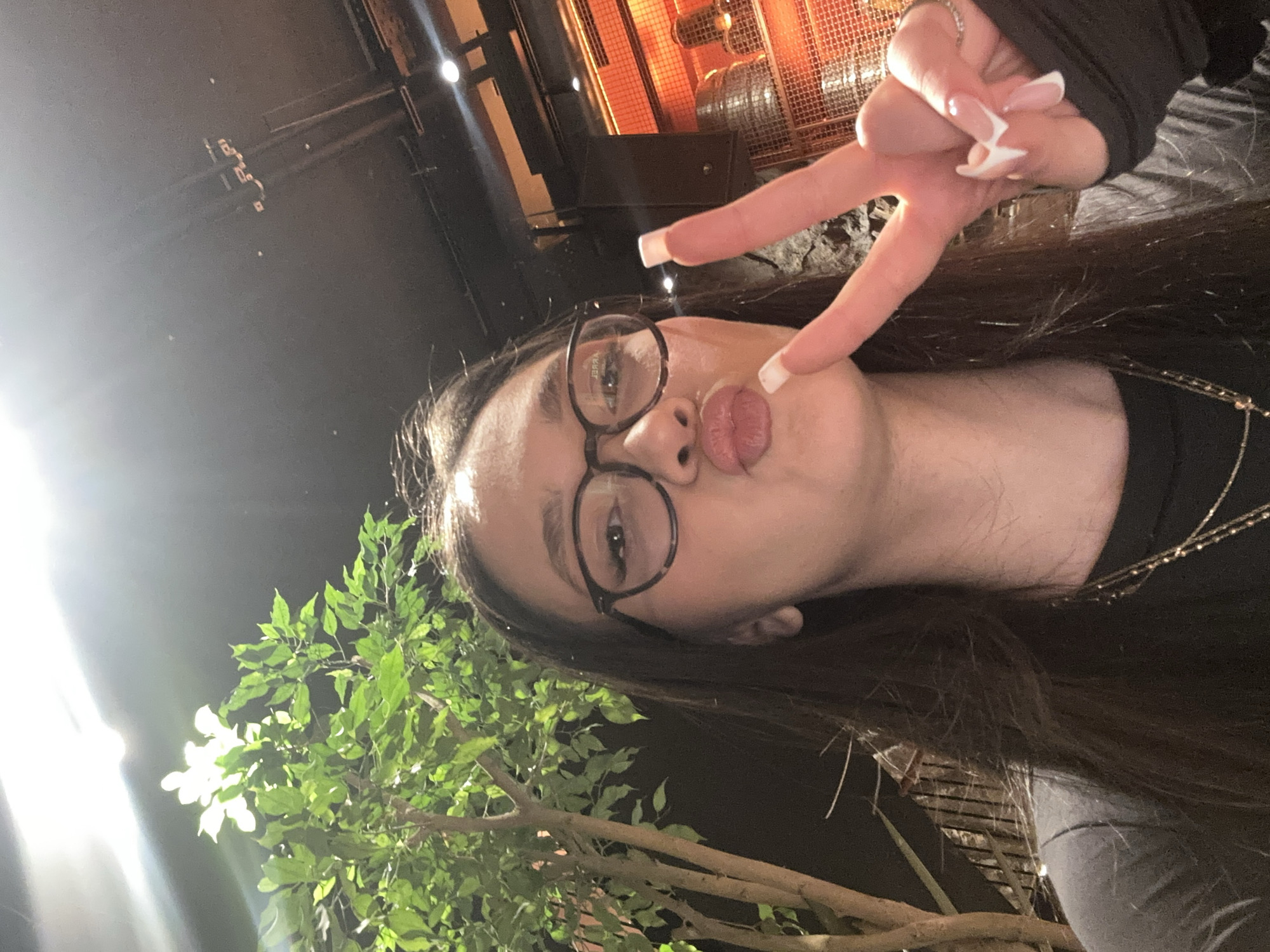 A woman with brown hair and glasses makes a kissing face while giving a peace sign. She is outside at night, near green foliage, likely in Yerevan, Armenia. Overhead lighting and architectural features are visible.