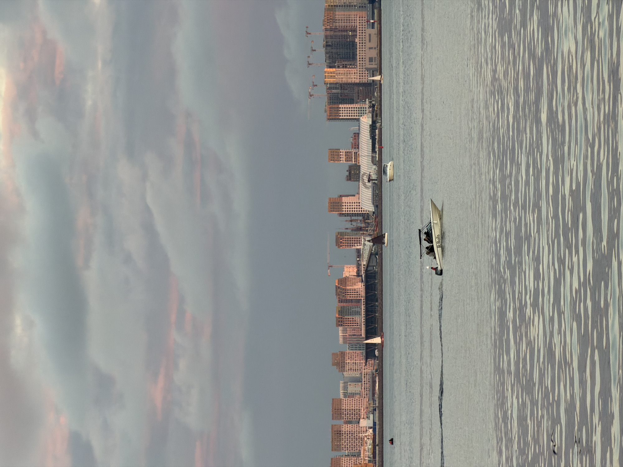 This is a photograph of a body of water with a city skyline in the background. The sky is cloudy with streaks of pink and gray. The water is calm and reflects the sky. In the foreground, a small white motorboat with several people aboard is moving from left to right, leaving a wake behind it. The boat has the word "Rolls" visible on its side. In the middle ground, there are several sailboats and another small motorboat. The city skyline consists of numerous modern buildings, some of which appear to be under construction, with several cranes visible. The buildings are a mix of light brown and white, with many windows. The overall impression is one of a calm day on the water with a developing urban landscape. The location is identified as St. Petersburg, Russia.