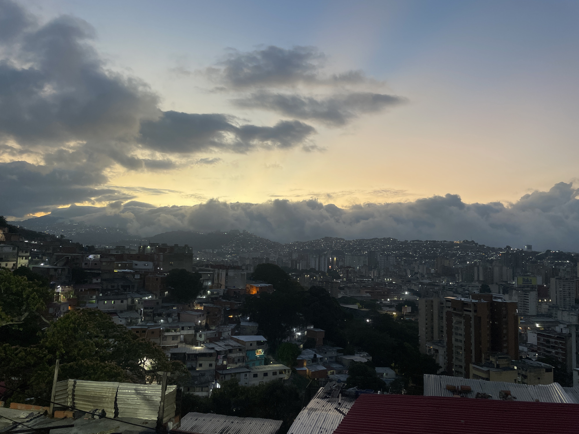 Here is a detailed description of the image:
The image presents a high-angle view of a cityscape at dusk or dawn. The location is Caracas, Venezuela. The scene is dominated by densely packed buildings and houses, many of which appear to be in a state of disrepair. These residential areas stretch across uneven terrain and up hillsides. The buildings range in size from small, single-story dwellings to taller apartment blocks.
In the foreground, there are several corrugated iron rooftops, some painted red. These roofs give way to a cluttered landscape of homes and structures nestled amongst trees. Further back, the city transitions into more densely built-up areas, with high-rise buildings visible in the distance.
The sky is partially covered by clouds. Closer to the horizon, there is a band of golden-yellow light, suggesting either sunrise or sunset. Darker, heavier clouds are scattered across the sky, casting shadows over the city.
Small lights are illuminated across the urban area, adding a warm glow amidst the dimness. The overall impression is one of an urban environment transitioning from day to night, or vice versa, with a mix of residential and commercial buildings packed densely together.