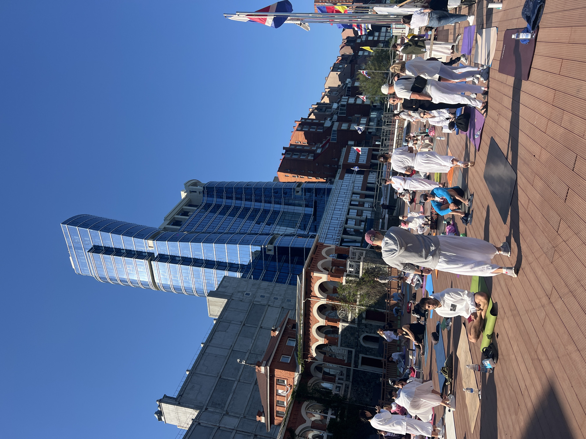 A wide, slightly low-angle, outdoor shot shows a yoga class in progress on a wooden boardwalk in Vladivostok, Russia, against a backdrop of modern and historic buildings under a clear blue sky.
The foreground features a group of people engaged in yoga on various colored yoga mats, with most wearing white clothing. The boardwalk is made of brown wooden planks.
Behind the yoga class, there's a mix of architectural styles. To the left, a modern, tall building with a curved, blue glass facade stands out. Adjacent to it is a grey, concrete building. To the right, a historic building with red brick arches is visible.
At the top of the frame, flags of various nations are displayed.