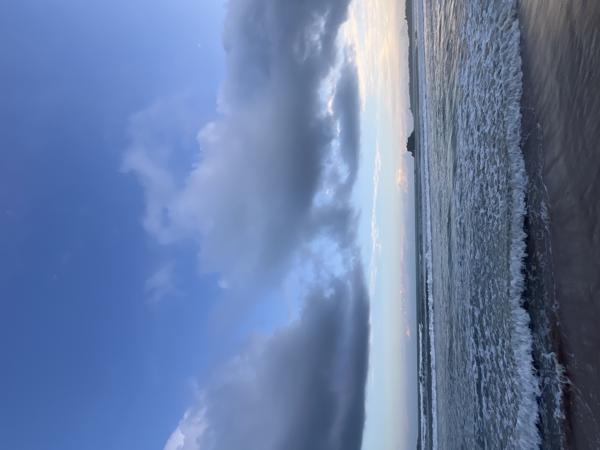 A wide-angle shot captures a serene beach scene in Peleana, Sri Lanka. The image is dominated by a vast, blue sky filled with scattered, moody clouds. Below, a calm ocean stretches towards the horizon, with gentle waves breaking on the sandy shore. A small, rocky island is visible in the distance. The lighting suggests it may be dawn or dusk, casting a soft glow over the scene. The sand in the foreground is wet from the receding tide.