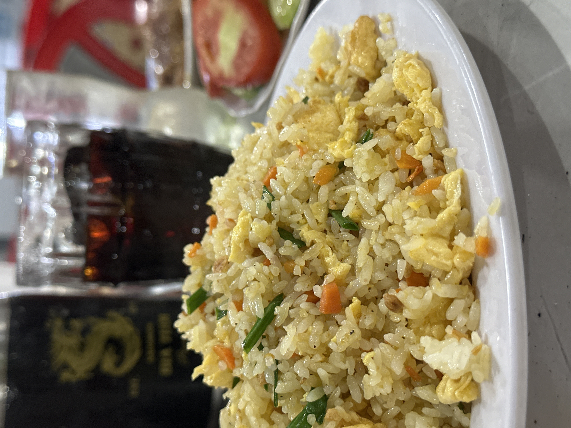 The image depicts a close-up of a prepared meal served on a light-colored table in an indoor dining setting. The scene is situated in Phu Thuy Ward, Vietnam.
In the foreground, a white, oval-shaped plate holds a generous mound of fried rice. The rice grains are light yellow to off-white, indicating a well-cooked and seasoned dish. Visible ingredients within the rice include irregular pieces of bright yellow scrambled egg, small diced orange carrots, and thinly chopped dark green scallions or spring onions. Some tiny dark specks, likely pepper or other seasoning, are scattered among the grains. The rice appears to be relatively dry, characteristic of well-prepared fried rice.
In the blurred midground, to the left of the fried rice, are two distinct beverage containers. Closest to the rice is a clear glass containing a dark brown liquid, consistent with a carbonated soft drink or iced tea. Reflections of light are visible on its surface. Further left, and more out of focus, is a dark-colored, possibly glass, bottle. A golden emblem, resembling a stylized dragon or a letter "S," is visible on its label, along with some indistinct text below it.
To the right of the fried rice plate, in the heavily blurred background, another white plate is visible, containing what appears to be a side salad or garnish. Slices of red tomato are discernible, with one prominent slice showing a lighter, possibly yellowish, central core. Green slices, consistent with cucumber, are also partially visible. Beyond this, a heavily blurred red object, possibly part of a restaurant fixture like a menu holder or napkin dispenser, can be faintly seen.
The table surface beneath the plates is a light grey or white, with a subtle textured or speckled pattern. The overall lighting is bright, suggesting an indoor artificial light source. No people are visible in the frame, and no specific actions or interactions are depicted, beyond the static presentation of the meal.