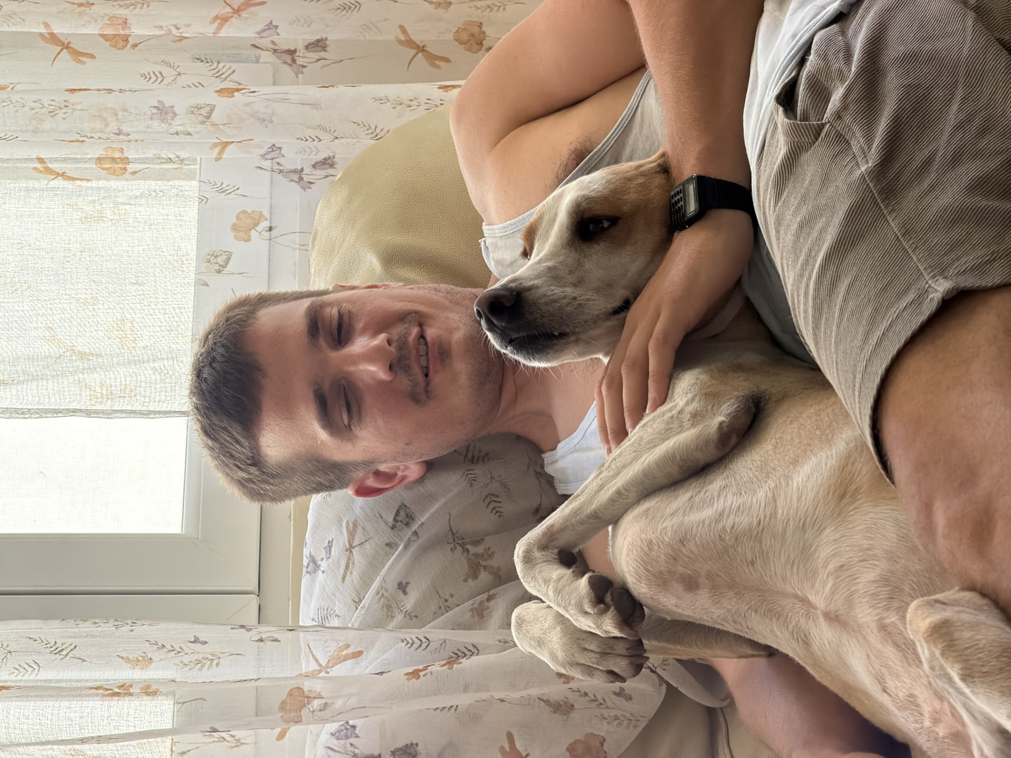 The scene is set indoors in Budva, Montenegro, depicting an adult male and a domestic dog in a relaxed posture on a sofa or cushioned seating. Natural light enters from a window positioned behind the subjects, which is partially obscured by two panels of sheer, light-colored curtains. The curtains are patterned with autumnal flora, including leaves and flowers in shades of orange, purple, and brown, and insects resembling dragonflies. A decorative pillow with similar botanical and insect patterns is visible behind the male.
The male, appearing to be in his late 20s to early 30s, has short brown hair and light facial stubble. He is reclined, with his eyes closed or mostly closed, and a gentle smile on his face. He wears a light-colored sleeveless tank top and striped light brown or beige shorts. A black digital watch with a calculator-style keypad is worn on his left wrist.
The dog, a light fawn or cream-colored mixed breed with white underparts, is positioned on its back, resting on the man's lap and chest. Its head is turned upwards, resting on the man's left shoulder and chest area, with its eyes open. Its front paws are bent, resting on its own body near its belly. The man's left hand is resting on the dog's chest/neck area, maintaining physical contact. The overall posture of both subjects indicates a relaxed and close interaction.