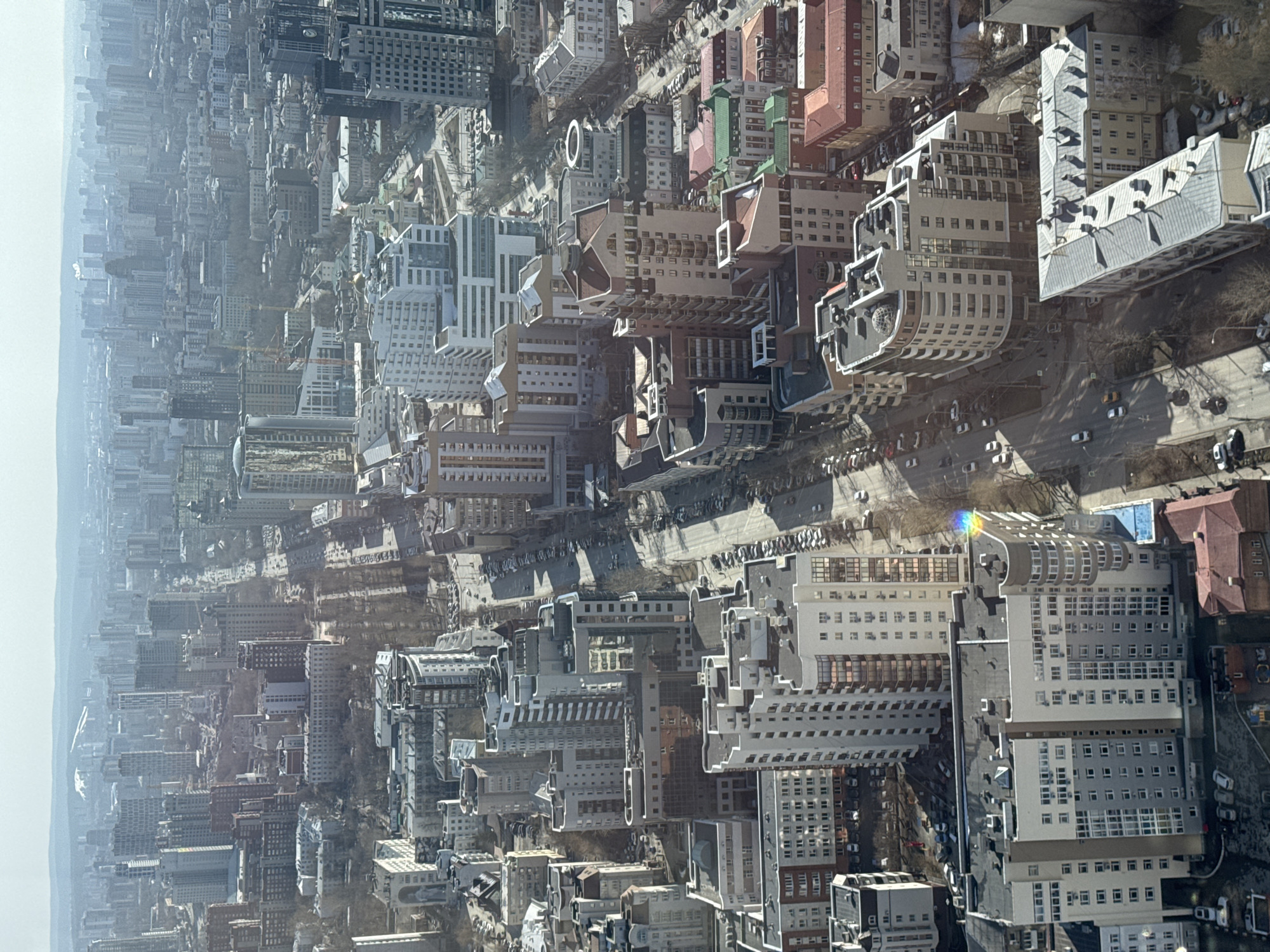 The image is an aerial shot of a densely populated urban area, seemingly of the city of Unknown. The foreground and midground are dominated by closely spaced buildings of varying heights and architectural styles. The rooftops display a mix of colors and textures, from flat concrete to pitched tile. A network of streets runs between the buildings, showing some vehicular traffic. In the background, a light haze obscures distant structures, creating a sense of depth. The light suggests it is daytime.