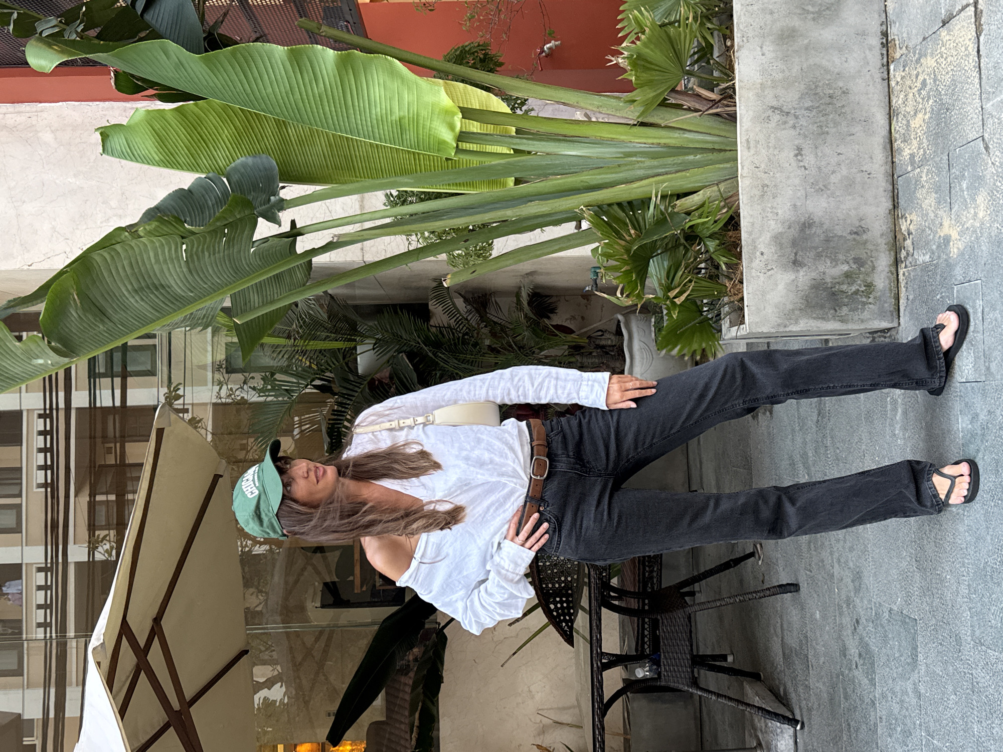 A woman stands outdoors on a paved area, likely a sidewalk or patio. She is wearing a green baseball cap, a white long-sleeved shirt, a brown belt, black jeans, and black sandals. She has long hair and a casual posture. To her left, there is an outdoor seating area with a white umbrella and rattan chairs. Lush green tropical foliage, including large banana leaves, is visible behind and to the side of her, suggesting a tropical climate. A building with windows is in the background. Based on the style of buildings and vegetation, it is possible that this photo was taken in the city of Đà Nẵng, Vietnam. The lighting suggests it is daytime.