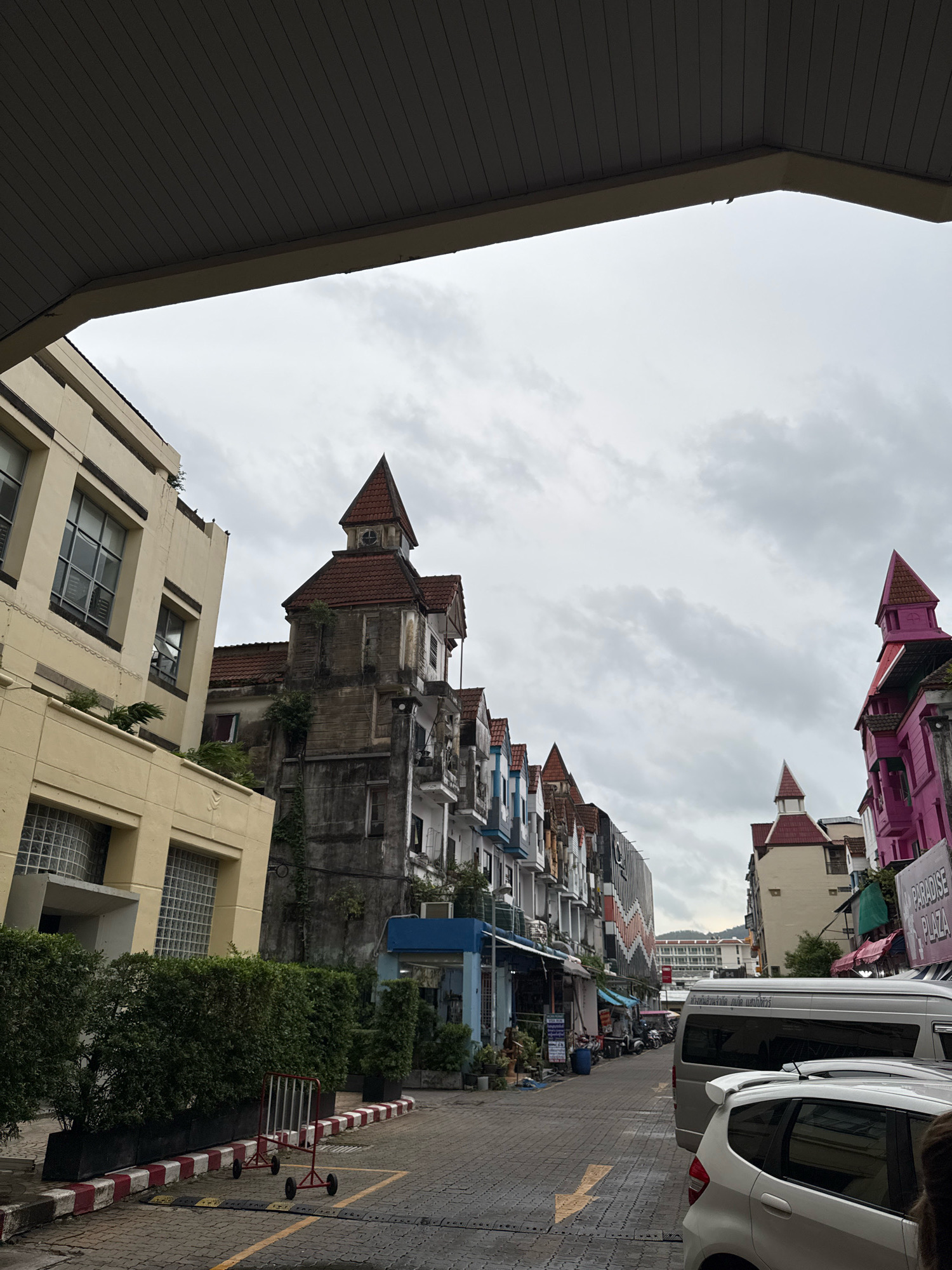 The image captures a street scene in Patong, Thailand, characterized by a mix of architectural styles and a cloudy sky. On the left, a cream-colored building with glass block windows and modern fenestration stands prominently. Adjacent to it is a weathered, dark stone building with a red-tiled turret, overgrown with vines. This building transitions into a row of narrow, colorful structures with similar red-tiled roofs, painted in shades of blue, grey, and white. Further down the street, a vibrant pink building with a matching turret stands out.
The street itself is paved with interlocking bricks. A white van and a white hatchback are parked on the right side of the street. In the foreground, a low hedge with neatly trimmed bushes is bordered by a curb with red and white markings. A portable metal barrier on wheels is situated on the side of the road. Various shops and businesses appear to line the street, indicated by signs and awnings, though many are not clearly legible. The overall atmosphere suggests a commercial area in a tropical climate, under an overcast sky.