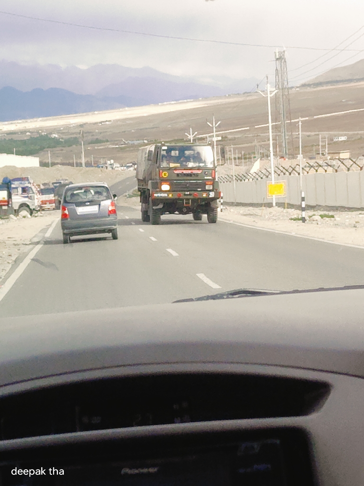 The image shows a road scene in Spituk, India, with a military truck in the foreground, followed by a gray car. The road is asphalt with white markings. In the background, there are mountains and a power line tower. The scene appears to be a typical day on a highway in a mountainous region.
