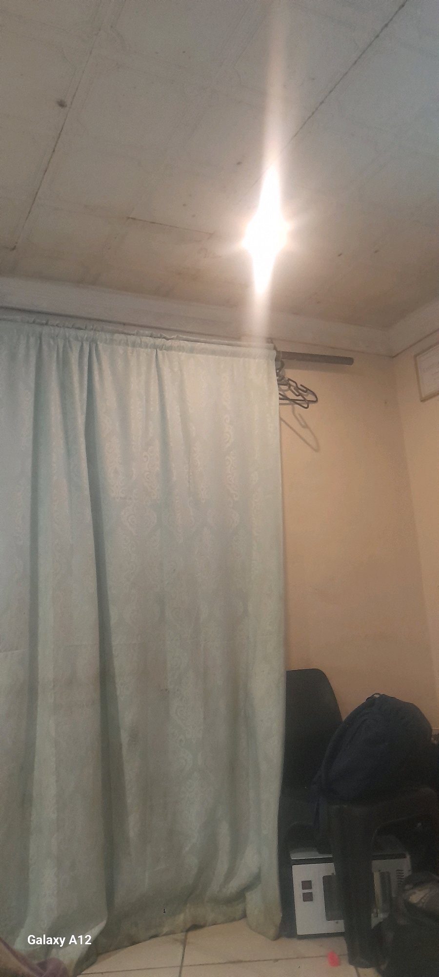 The image shows an interior room with a white ceiling and pale yellow walls. A pale blue curtain with a subtle damask pattern covers a large portion of the left side of the frame. Above the curtain, a metal curtain rod is visible, with several wire hangers hanging from it. A bright light source, possibly a bulb, is shining from the ceiling on the right side of the frame, creating a strong glare. In the lower right corner, a dark chair is partially visible, with a dark blue bag on top of it. In front of the chair, a white electronic device with red indicator lights is on the floor. The floor appears to be tiled. The lower left corner of the image displays "Galaxy A12". Based on the information provided, this scene is located in Blue Downs, South Africa.