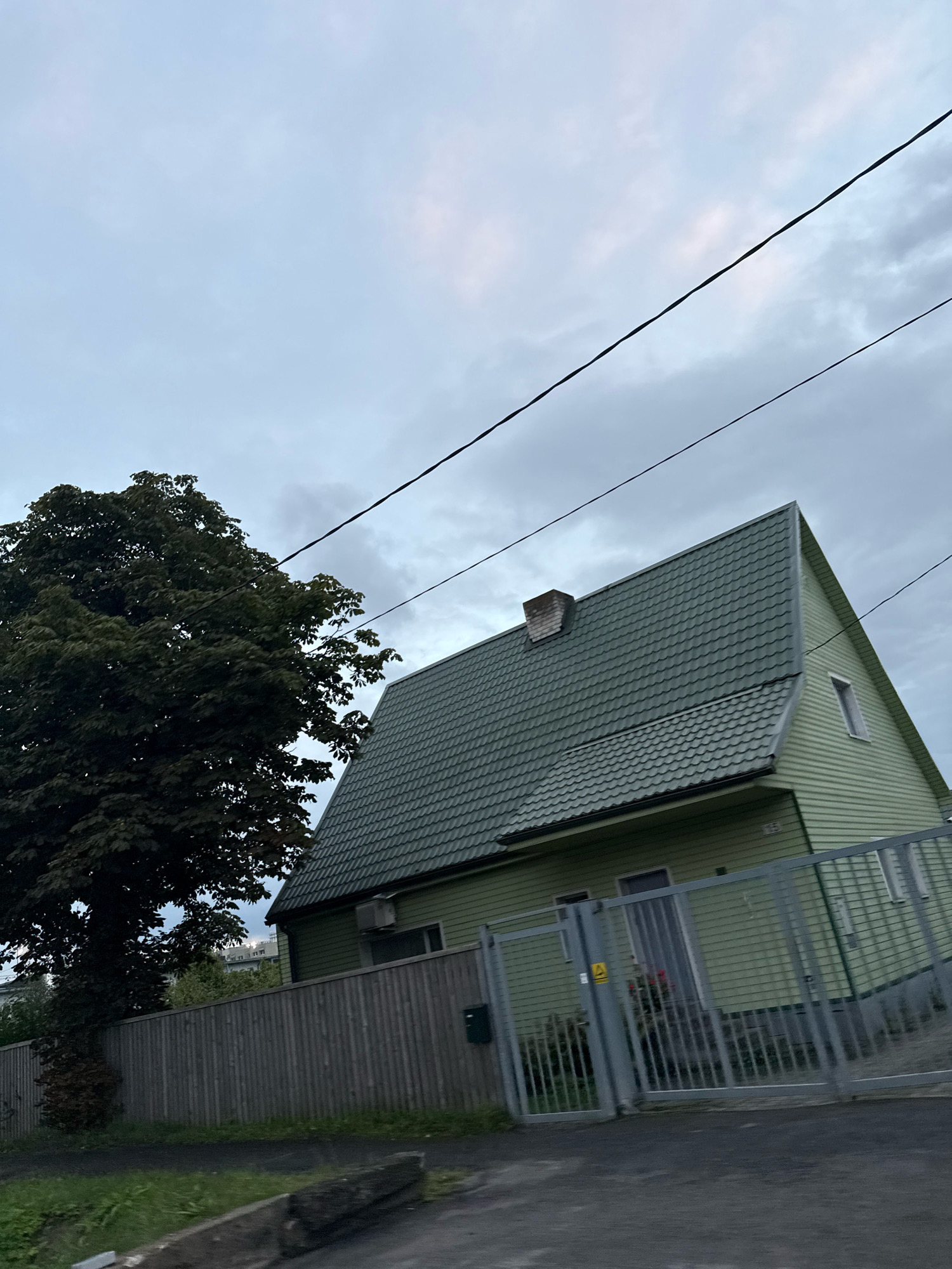 The image captures a side view of a light green, two-story house with a dark green, shingled pitched roof. A brick chimney is visible on the roof. The house has several windows, including a small, narrow window on the upper right gable. An air conditioning unit is mounted on the exterior wall below the roofline. The house is bordered by a gray, vertical plank fence and a tall, gray metal gate. A mature tree with dense dark green foliage stands to the left of the house, partially obscuring it. Overhead, two black power lines stretch across the cloudy, pale gray sky. The scene appears to be a residential area.
