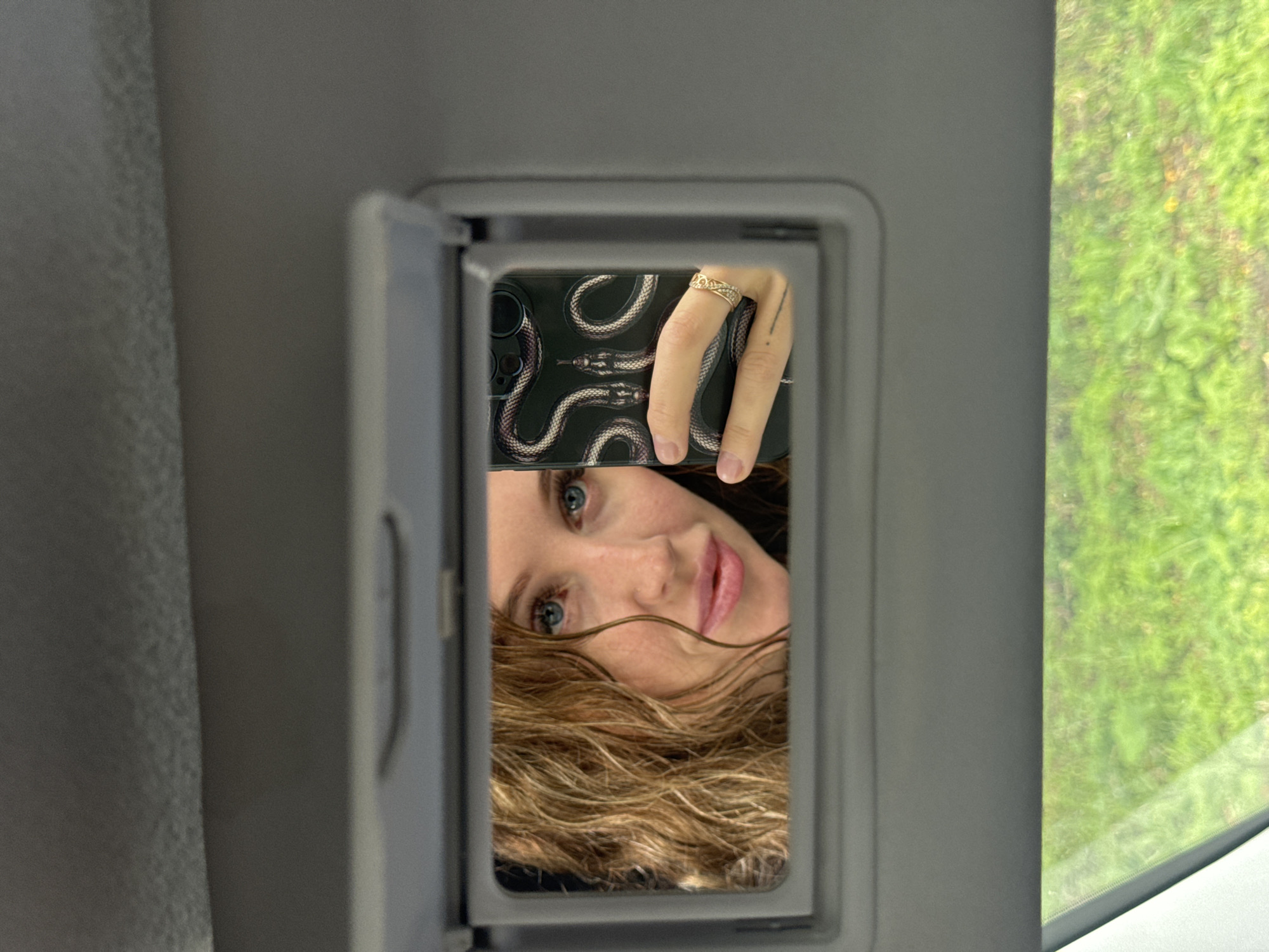 A woman with long, curly blonde hair and blue eyes takes a selfie in a car's vanity mirror. She is holding a black smartphone with a snake-patterned case. She is wearing a ring on her left hand. The background is blurred green grass. The location is not specified in Chaikovsky, Russia.