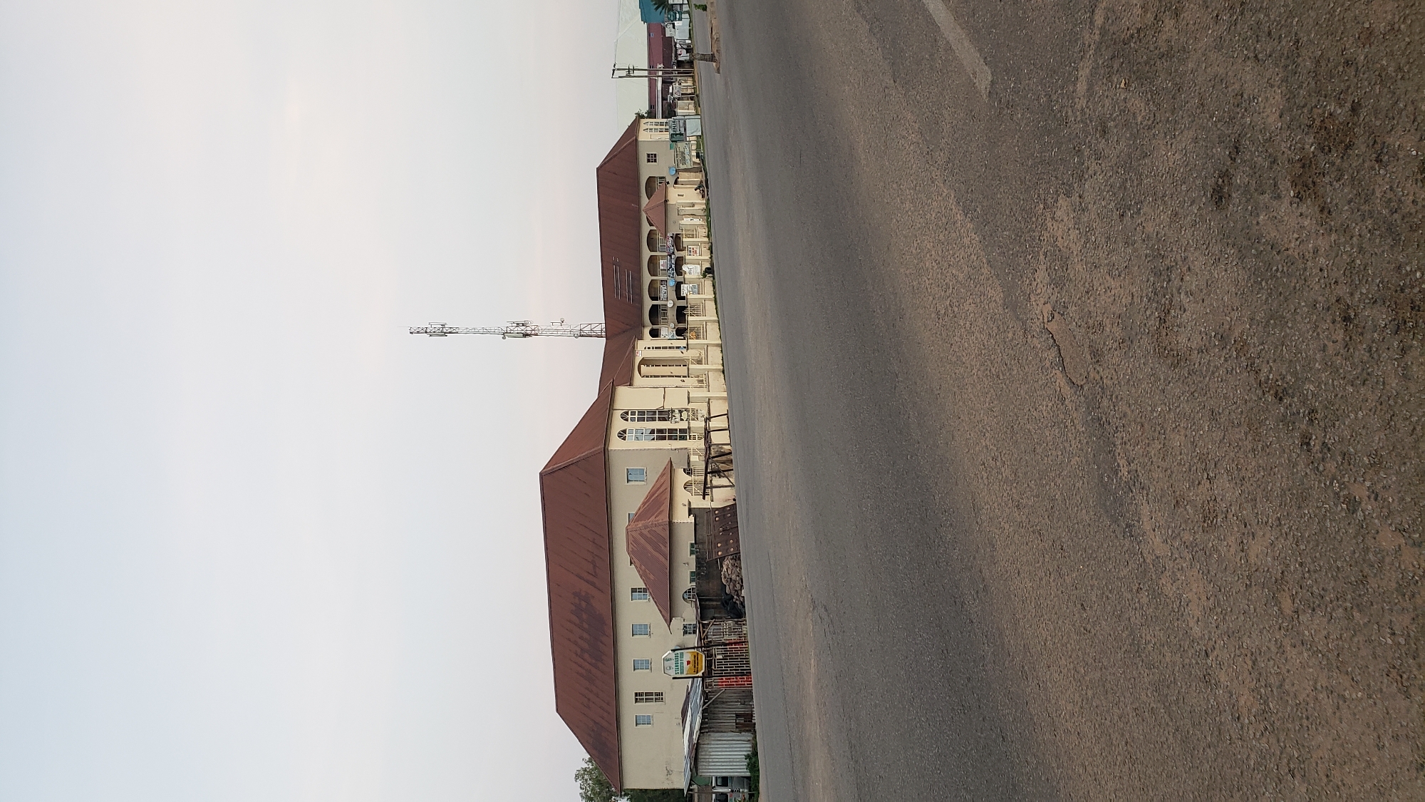 The image depicts a street scene in Jalingo, Nigeria. The primary subjects are buildings and a road. The buildings appear to be commercial or residential, with multiple stories, balconies, and reddish-brown roofs. One building has a prominent telecommunications tower adjacent to it. The road takes up a significant portion of the frame, appearing to be asphalt with a white line marking.
The scene seems to be set during the day, likely late afternoon or early morning, given the soft light and the long shadows cast by the buildings. The sky is overcast or hazy, suggesting a neutral weather condition. There are no people visible in the immediate foreground or mid-ground of the image, and no specific activity is discernible beyond the static presence of the buildings and the road.
Several details can be observed on the buildings. Signs are visible, although the text is largely unreadable due to resolution and distance. One sign appears to be yellow and green. There are also some stacks of materials or goods placed near one of the buildings, possibly indicating commercial activity or storage. The overall impression is one of an urban or semi-urban environment in Nigeria.