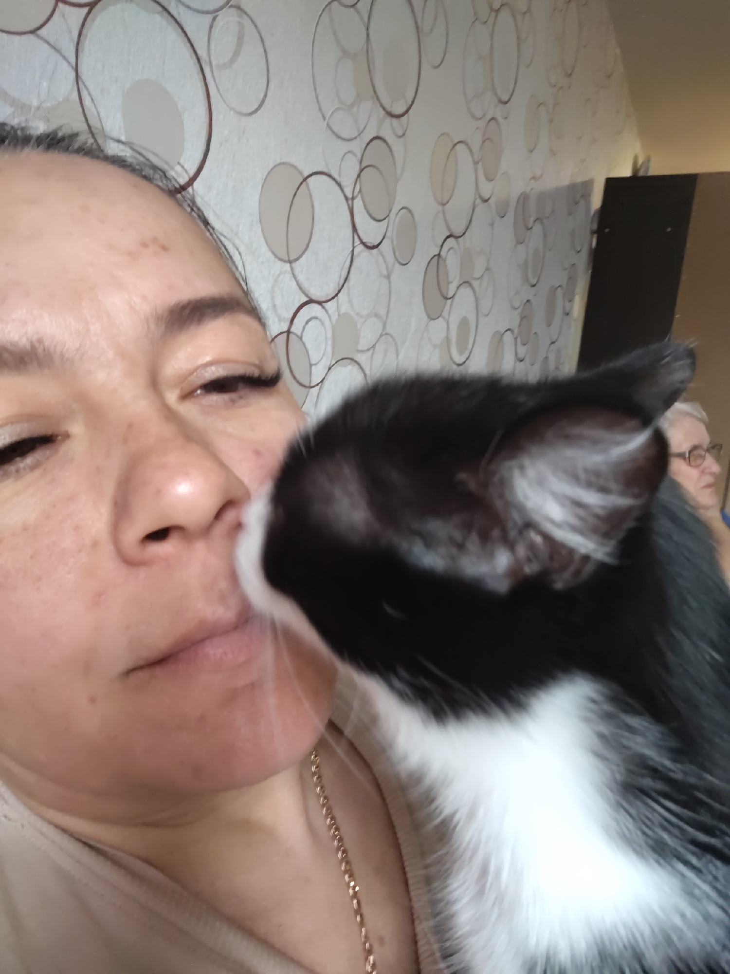 A medium shot depicts a woman's face interacting with a black and white cat, indoors. The woman's face is close to the camera, with her left eye partially closed, and her nose and mouth are positioned near the cat. Her skin tone is light, with freckles visible on her cheeks and nose. The cat's head is angled towards her face, with its nose appearing to touch her face. The cat has black fur on its head and back, with white fur covering its chest area.
In the background, a wall covered in a patterned wallpaper of overlapping circles in varying shades of brown and white. To the right of the woman, partially obscured by the cat's head, is an older woman wearing glasses, further back in the scene. A dark rectangular object is seen behind the woman, possibly a piece of furniture.