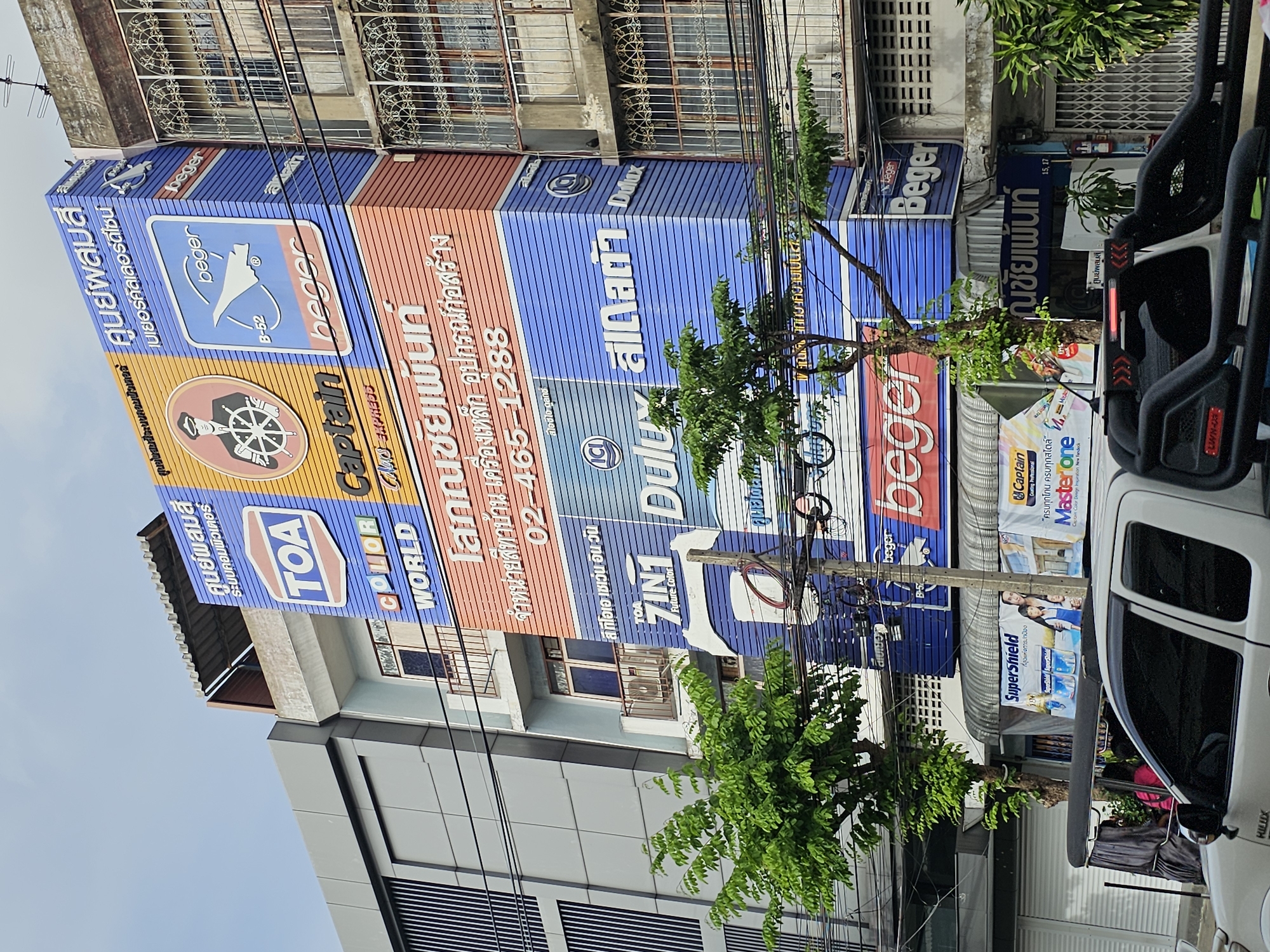 The image shows a multi-story building in Bangkok, Thailand, with numerous advertisements for paints and construction materials displayed prominently on its facade. The building appears to be a commercial establishment, possibly a showroom or a paint supplier. The advertisements feature brands like TOA, Captain, Dulux, and Beger. The text on the advertisements is primarily in Thai, with some English brand names visible.
In the foreground, a white pickup truck with a black roll bar is parked on the street. A three-wheeled motorized vehicle, commonly known as a tuk-tuk, is partially visible behind the pickup truck, with passengers seated inside. The street is lined with buildings, power lines, and trees. The sky is overcast. The overall scene depicts a typical street view in a bustling urban environment of Bangkok, characterized by its vibrant commercial signage and active street life.
