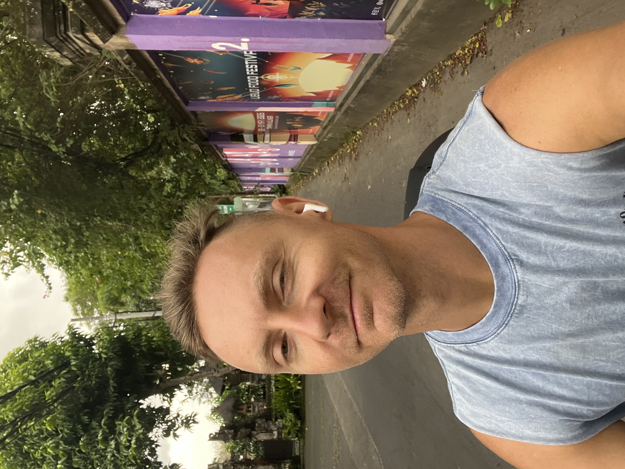 A head-and-shoulders selfie shows a fair-skinned man with short blond hair, wearing a blue sleeveless shirt and a white wireless earpiece. He is on a paved road next to a purple concrete wall decorated with colorful posters. The posters advertise the Ubud Food Festival with dates in May 2023. Beyond the wall, lush green trees and tropical foliage suggest the scene is in a park area or street in Ubud District, Indonesia.
