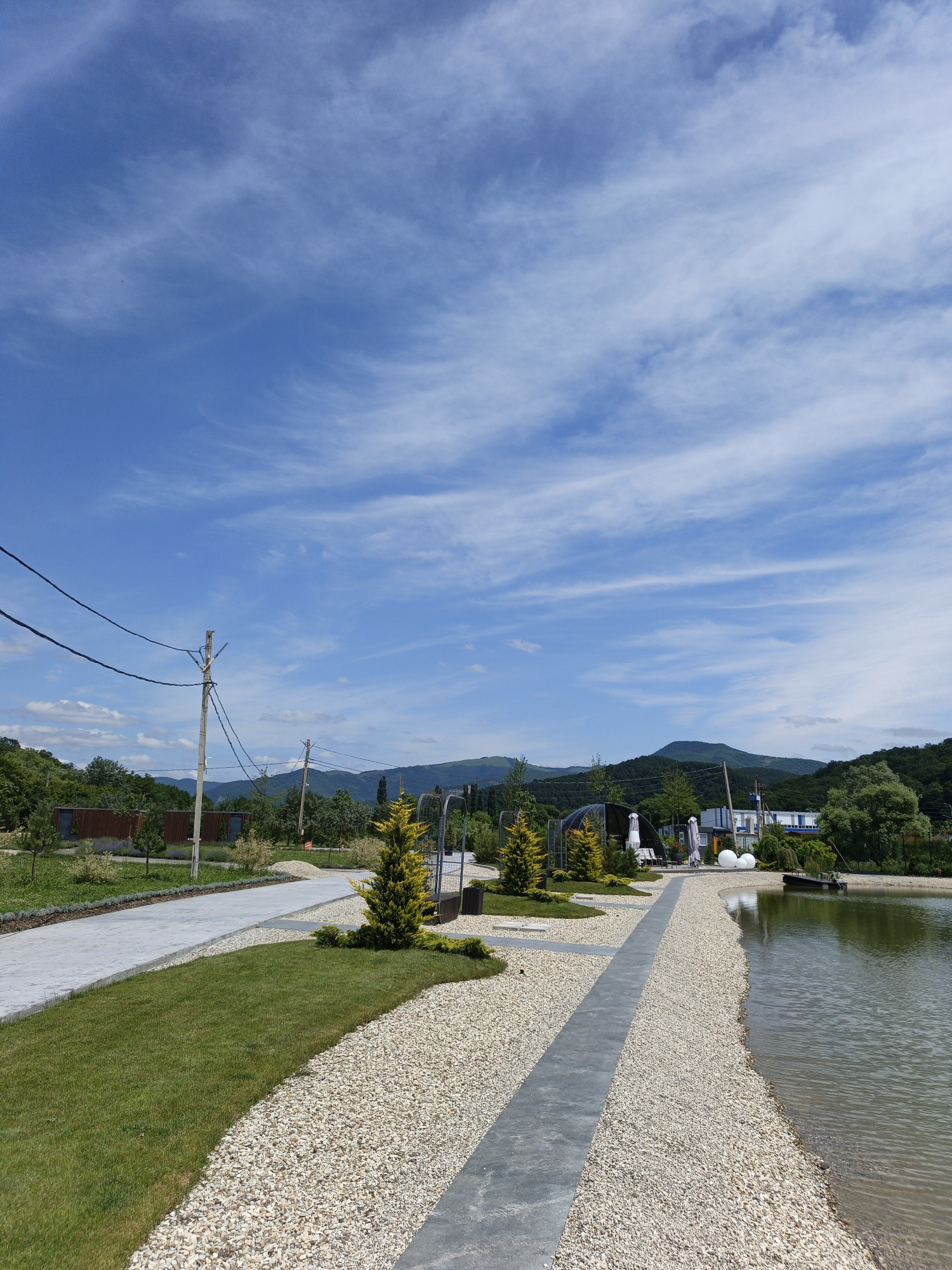 This is a vertical, eye-level shot of an outdoor recreational area with a lake, a paved walkway, and a grassy area. The scene appears to be a park or resort, likely in Pshada, Russia, given the location context.
The sky is a vibrant blue with wispy white clouds, indicating it is daytime and the weather is clear and pleasant. A power pole with several wires stretches across the top left of the frame. In the background, lush green hills and mountains rise, suggesting a natural, possibly rural or semi-rural, setting.
The foreground is dominated by a path made of light-colored gravel and concrete slabs, curving along the edge of a calm body of water. A small boat is docked on the lake. Adjacent to the path is a well-maintained lawn. Scattered within the park area are several small, conical evergreen trees and some modern-looking structures, including a semi-circular, transparent dome that houses lounge chairs and umbrellas. Two large, white spherical objects are visible near the dome, possibly decorative elements. There are no people visible in the frame. The overall impression is one of tranquility and a well-designed leisure space.