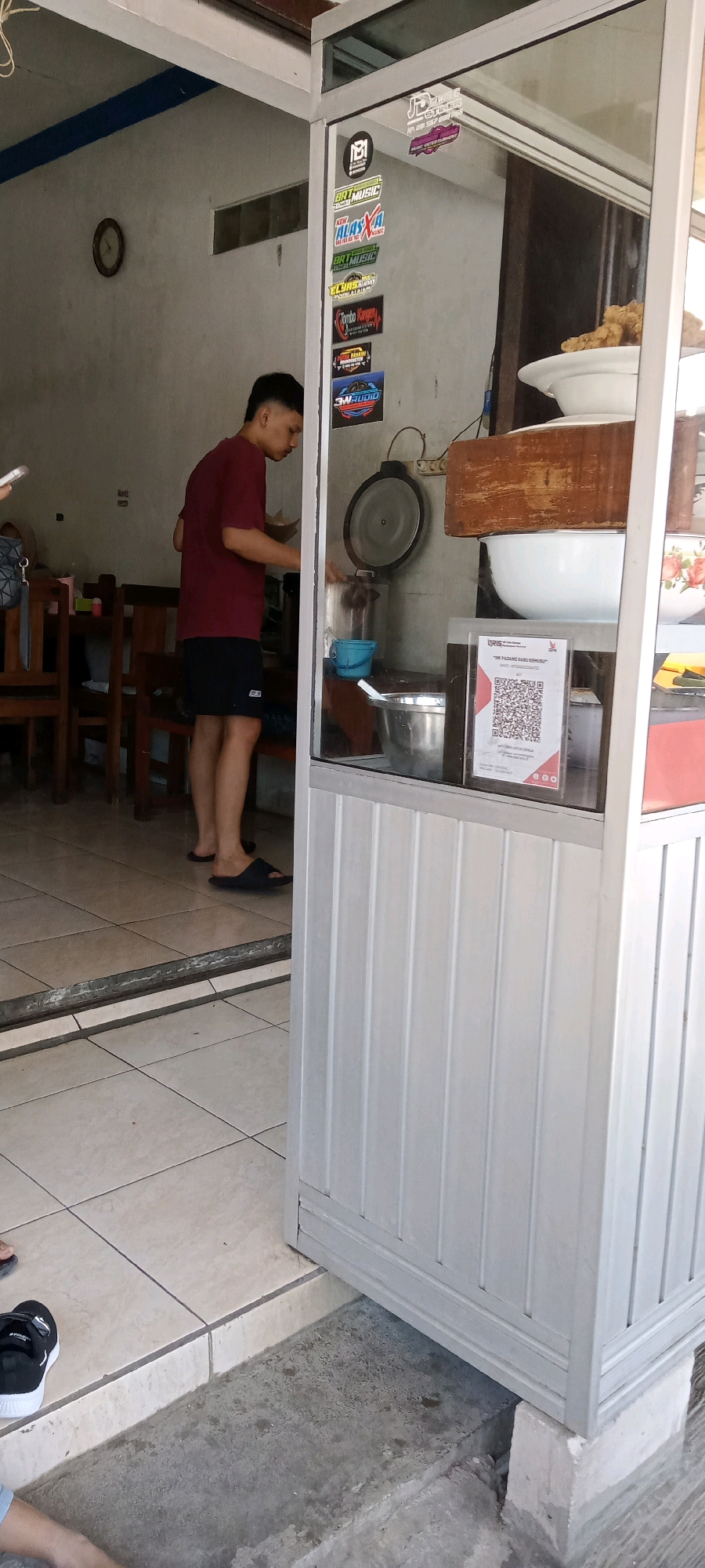 In the city of Desa Karangmojo, Indonesia, a young man wearing a maroon t-shirt and black shorts stands in what appears to be a food stall or small eatery. He is positioned near a counter with a glass enclosure, behind which various food items and cooking utensils are visible. On the counter, there is a large metal pot with a lid, a smaller blue bucket, and a stainless steel bowl. A QR code is displayed on the glass, presumably for payment or ordering. Behind the young man, a wall with a circular clock and various stickers can be seen. Wooden tables and chairs are arranged in the background, suggesting a dining area. The floor is tiled. The overall scene depicts a typical day at a local Indonesian food establishment.