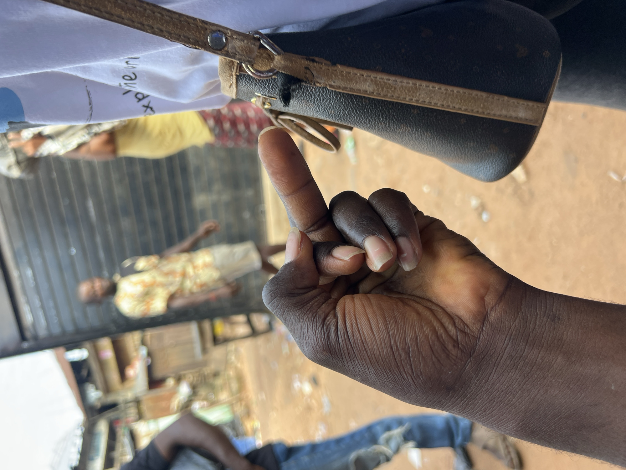 An image taken from a low angle shows a dark-skinned hand in the foreground with the middle finger extended. The hand appears to be positioned in front of a dark brown and tan patterned handbag. In the background, there are blurred figures of people in an outdoor setting with a dirt ground. One person in the background wears a yellow and white patterned shirt and light-colored pants, and another wears a yellow shirt and patterned pants. The environment suggests a street scene in a market or a similar public space. The location is identified as Nkpor, Nigeria.