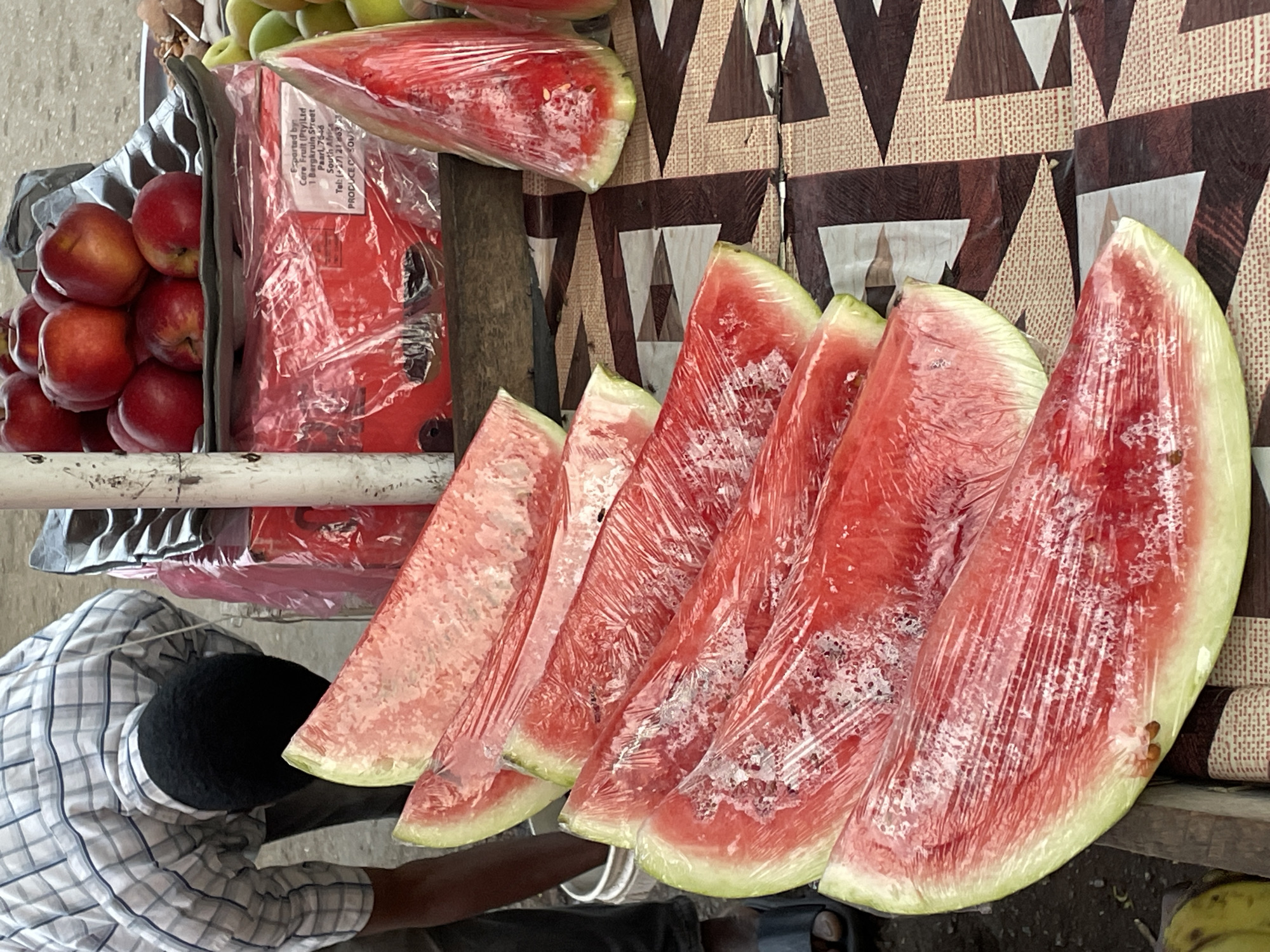 The image shows a vendor at a street market in Jos, Nigeria, selling various fruits. Red apples are stacked in a container, and slices of watermelon, individually wrapped in clear plastic, are arranged for sale. A whole watermelon wrapped in plastic is also visible. The vendor, dressed in a checkered shirt, stands next to the fruit stand. The background features a patterned fabric. A box with the text "Exported by Core Fruit (Pty) Ltd" is partially visible.