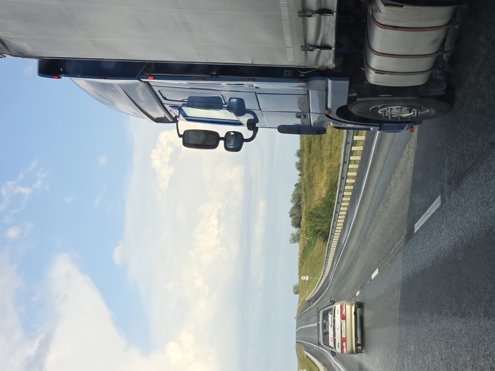 A blue semi-trailer truck travels on a highway in the city of Troitsk urban district, Russia. The truck is in the right lane, and a white sedan is in the left lane ahead of it. The highway is surrounded by grassy fields and trees. The sky is blue with some white clouds. The truck appears to be in motion. The sedan is also in motion, moving away from the camera. There are no people visible in the image. The environment appears to be rural or semi-rural.