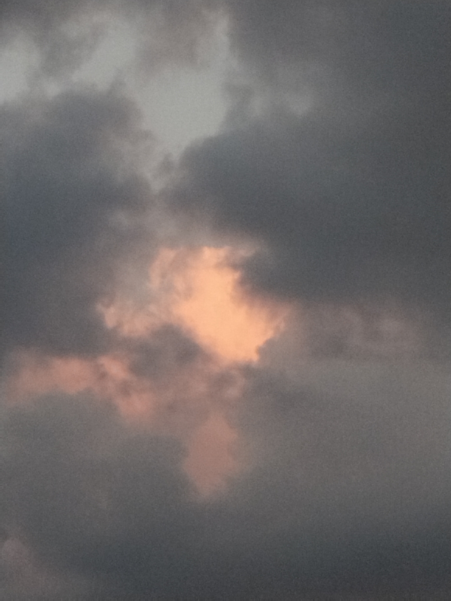 A close-up, abstract shot of the sky reveals dark, heavy clouds with a break in the center. Within this opening, lighter, peach-colored clouds are illuminated by an unseen light source, creating a contrast with the surrounding gloom. The image is out of focus and lacks any discernible landmarks or context, making it impossible to determine a specific location within any city.