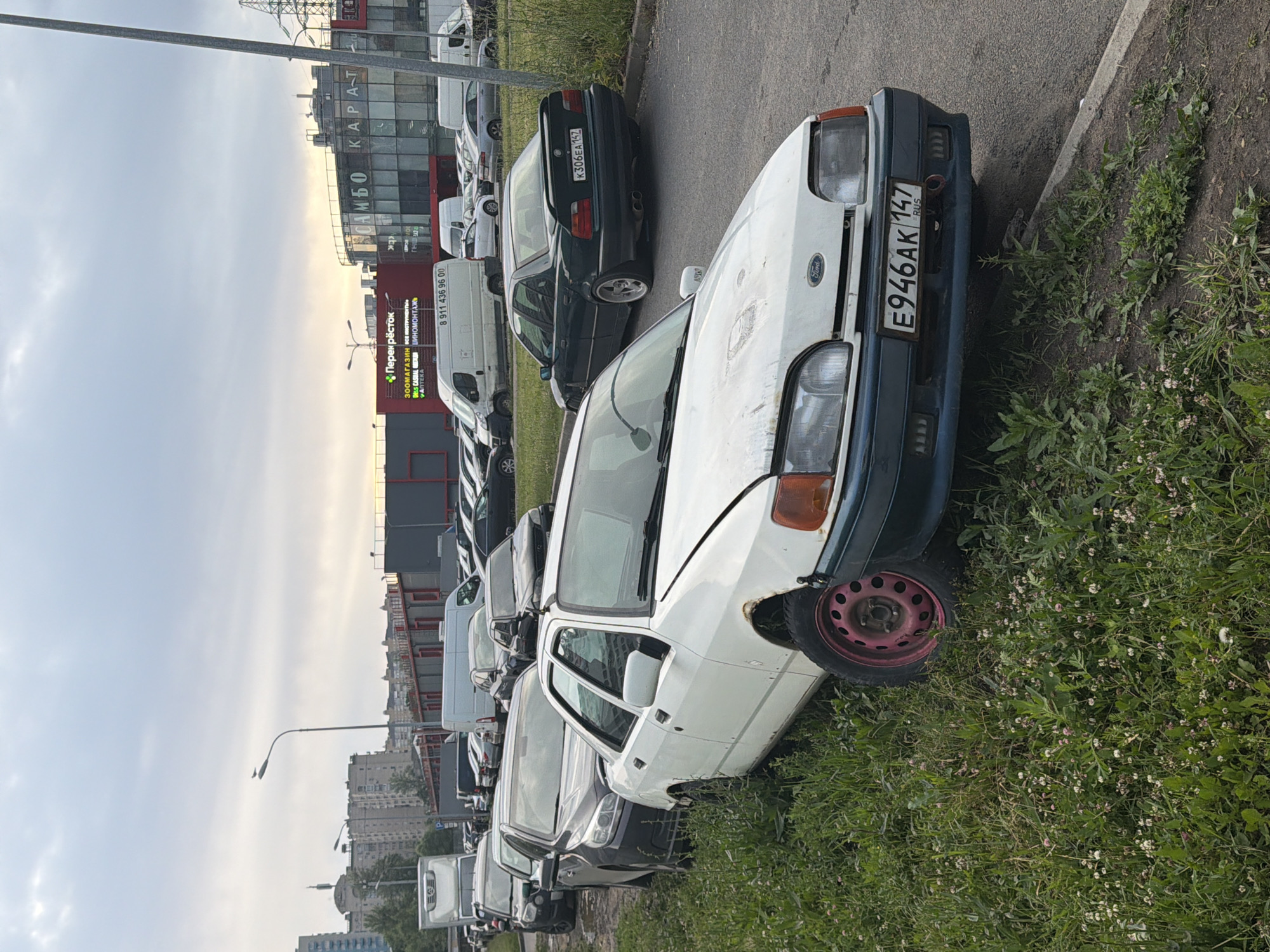 The image shows a row of parked cars on an asphalt road next to a grassy area. In the foreground, a white Ford Sierra is parked with its front partially on the grass. It has a pink wheel rim on the front passenger side. Further down the road, a dark green BMW sedan is parked, displaying the license plate K306EA 147. Behind these cars, other vehicles are visible, including vans and sedans. In the background, there are buildings and a cloudy sky. A street lamp is visible on the left side of the image, and a bird is perched on it. The scene appears to be an outdoor parking lot in an urban or suburban area.
