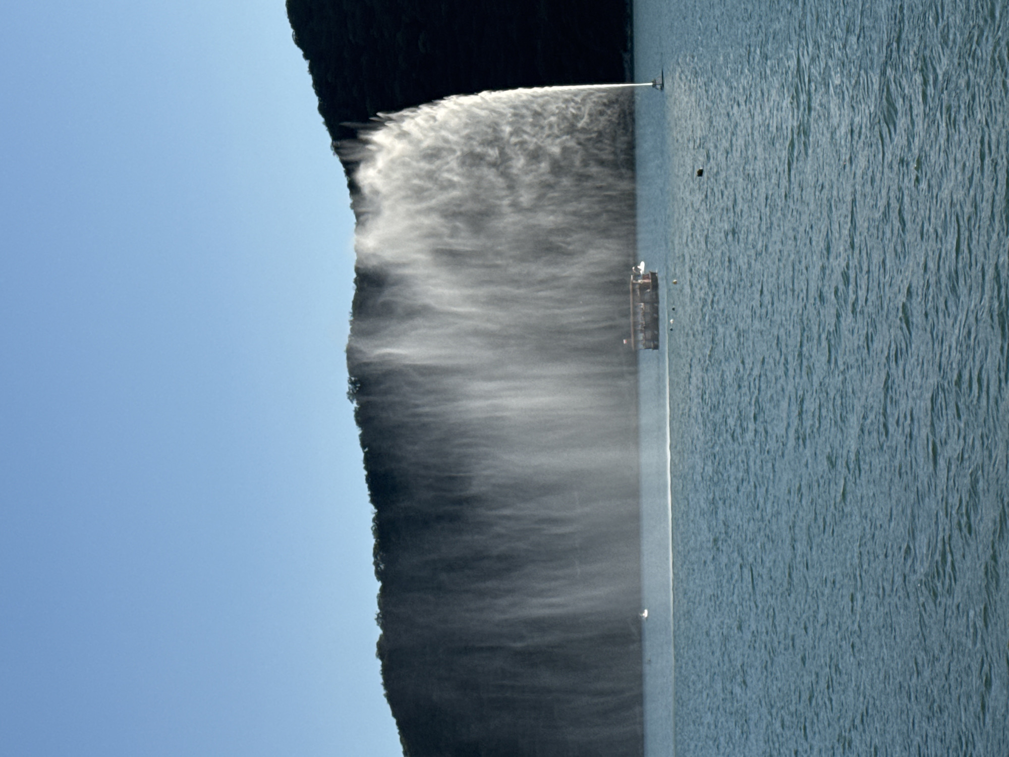 The image depicts a wide view of a body of water with a large fountain spraying water high into the air, forming a curtain-like effect against a dark, tree-covered hillside. The sky above is a clear, bright blue. In the foreground, the water is a deep blue-green, with ripples indicating a slight breeze.
Several boats are visible on the water. The most prominent is a long, multi-decked tour boat with a red roof, carrying passengers. To the right of the tour boat, and closer to the fountain, a smaller white boat or buoy-like structure is partially obscured by the spray. Further to the left, a very small white boat is seen in the distance.
The fountain itself appears to originate from a single point on the water and sprays upwards and outwards, creating a massive, opaque white wall of water that extends horizontally across the frame. The spray is illuminated by the sun, giving it a bright, luminous quality.
The hillside in the background is covered in dense, dark green foliage, suggesting a natural, perhaps mountainous or hilly, landscape. The overall scene captures a recreational setting, likely a tourist attraction, given the presence of the tour boat and the impressive man-made fountain.
The location of this impressive fountain is within the resort area of Abrau-Dyurso, Russia.