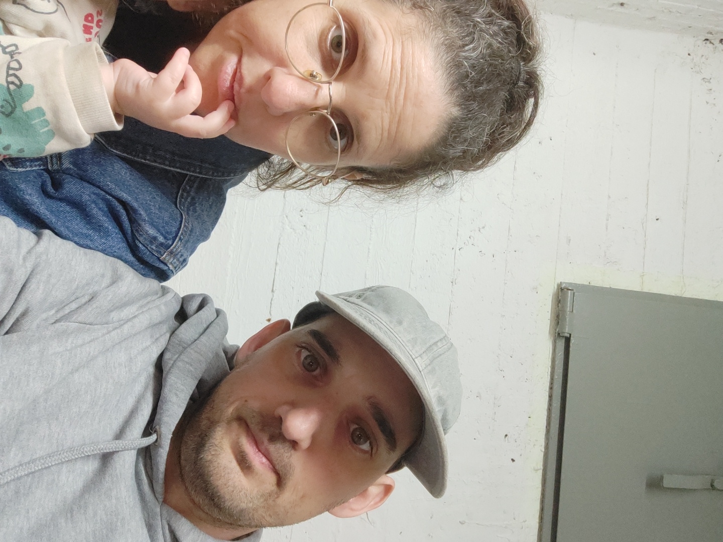 A selfie taken from a low angle shows a man, woman, and infant in an interior space with a concrete wall. The man is in the lower half of the frame wearing a light gray hooded sweatshirt and a light gray baseball cap. He has short brown hair and a beard. The woman, slightly above and to the left of the man, is wearing glasses and a denim jacket, with a child, who has a long-sleeved shirt with a pattern, leaning against her. A gray door and the concrete wall fill the right side of the image. Location cannot be determined from the provided image.