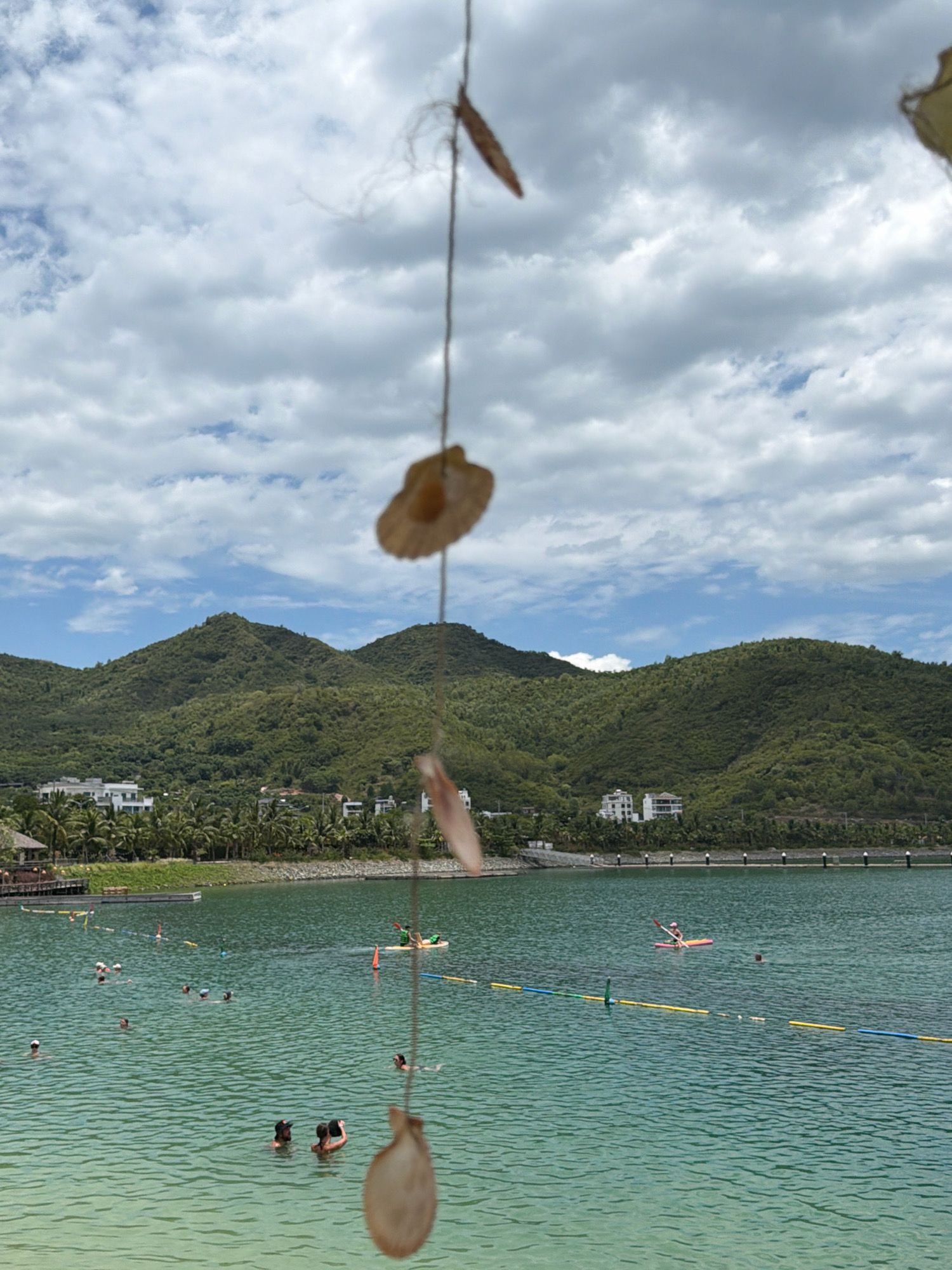 The image depicts a scenic coastal view in Nha Trang, Vietnam, featuring a tranquil bay bordered by lush green hills. The water is clear and turquoise, with several people swimming and engaging in water activities such as kayaking. The foreground is partially obscured by a decorative hanging made of shells, which adds a bohemian touch to the scene. In the background, buildings and palm trees line the shore, suggesting a resort or residential area. The sky is partly cloudy, with the sun casting a soft light on the landscape. The overall atmosphere is one of relaxation and natural beauty, typical of a tropical vacation destination.