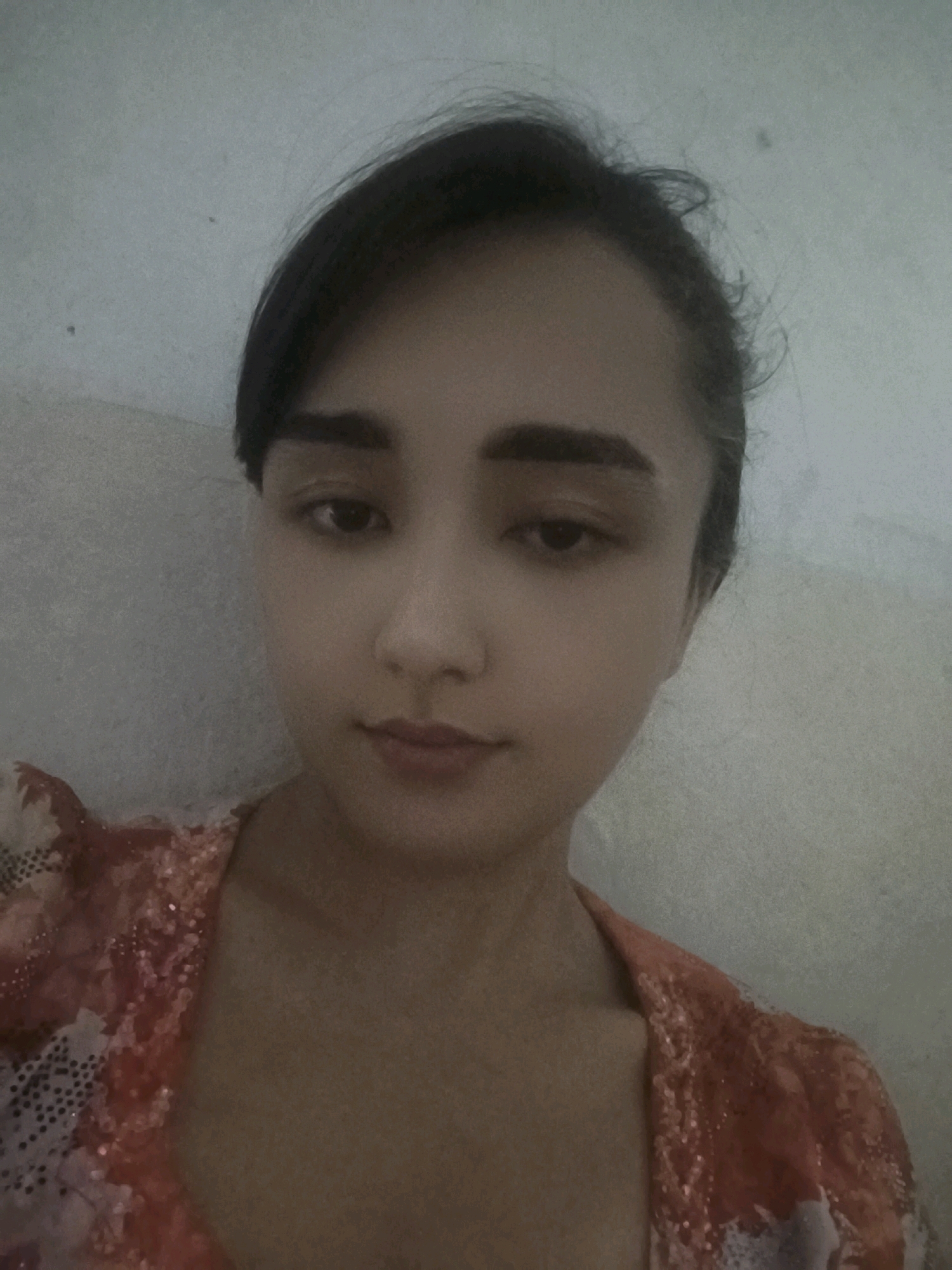 A frontal, medium-shot photograph captures a young woman from the chest up. She is positioned against a textured, pale grey wall, which appears to be indoors. Her dark hair is pulled back, with some wisps framing her forehead. She has distinct, dark eyebrows and her gaze is directed slightly downwards and to her left. Her makeup includes eyeshadow and lipstick, with a subtle hint of blush on her cheeks. She is wearing a garment with an orange, floral, and sequined pattern. The image quality is grainy and slightly out of focus, contributing to a soft, diffused lighting. The specific location in the city of Bokhtariyon, Tajikistan, is not discernible from the visual cues in the image.