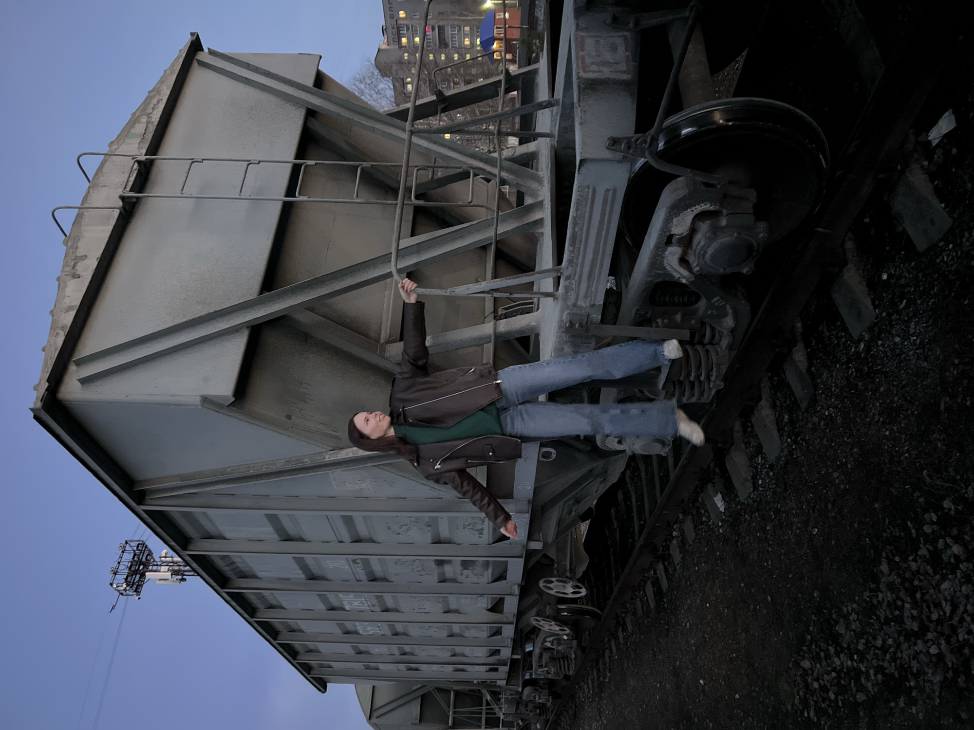 This rotated image captures a young woman posing horizontally against a large, grey freight train car, likely a hopper car, at dusk in Chaikovsky, Russia. The sky is a clear, muted blue, suggesting twilight, with some distant lights.
The woman has long brown hair and is dressed in a brown leather jacket, a green long-sleeved shirt, blue jeans, and white sneakers. She is suspended across the side of the train car, holding onto a metal ladder with her right hand, while her left arm hangs down. Her body appears to defy gravity due to the image's rotation, creating a visually striking pose. Her expression is neutral as she looks slightly upwards.
The train car is constructed from corrugated grey metal panels and sturdy structural beams, with faded numbers like "20" and "781E" visible on its side. Below the car, the dark gravel ballast of the train tracks and parts of the train's bogies and wheels are visible.
In the background, an urban landscape unfolds under the twilight sky. A distant building with multiple illuminated windows clearly displays Cyrillic text, including "БАНК" (BANK) and partially visible "ТОВАР" above it. Further afield, a tall, metallic lattice structure, possibly a cell tower or utility pole, rises against the sky. The scene combines industrial elements with a human subject in an artistic, gravity-defying composition.