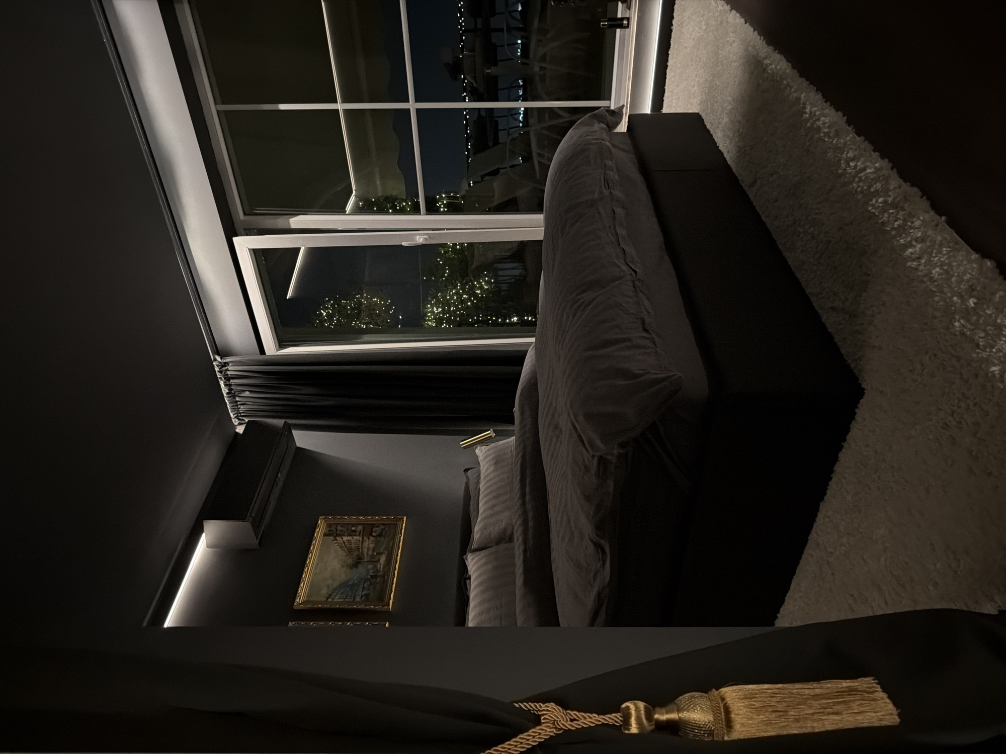A bedroom scene at night, featuring a large bed with dark grey bedding and a white shag rug. The room is dark, with ambient lighting from an air conditioning unit, accent lighting along the ceiling, and a bedside lamp. A framed painting hangs on the wall above the bed. Large sliding glass doors offer a view of a cityscape at night, with illuminated buildings and trees adorned with string lights. Outdoor furniture is visible on a balcony or patio. The location is described as a hotel room in Plovdiv, Bulgaria.