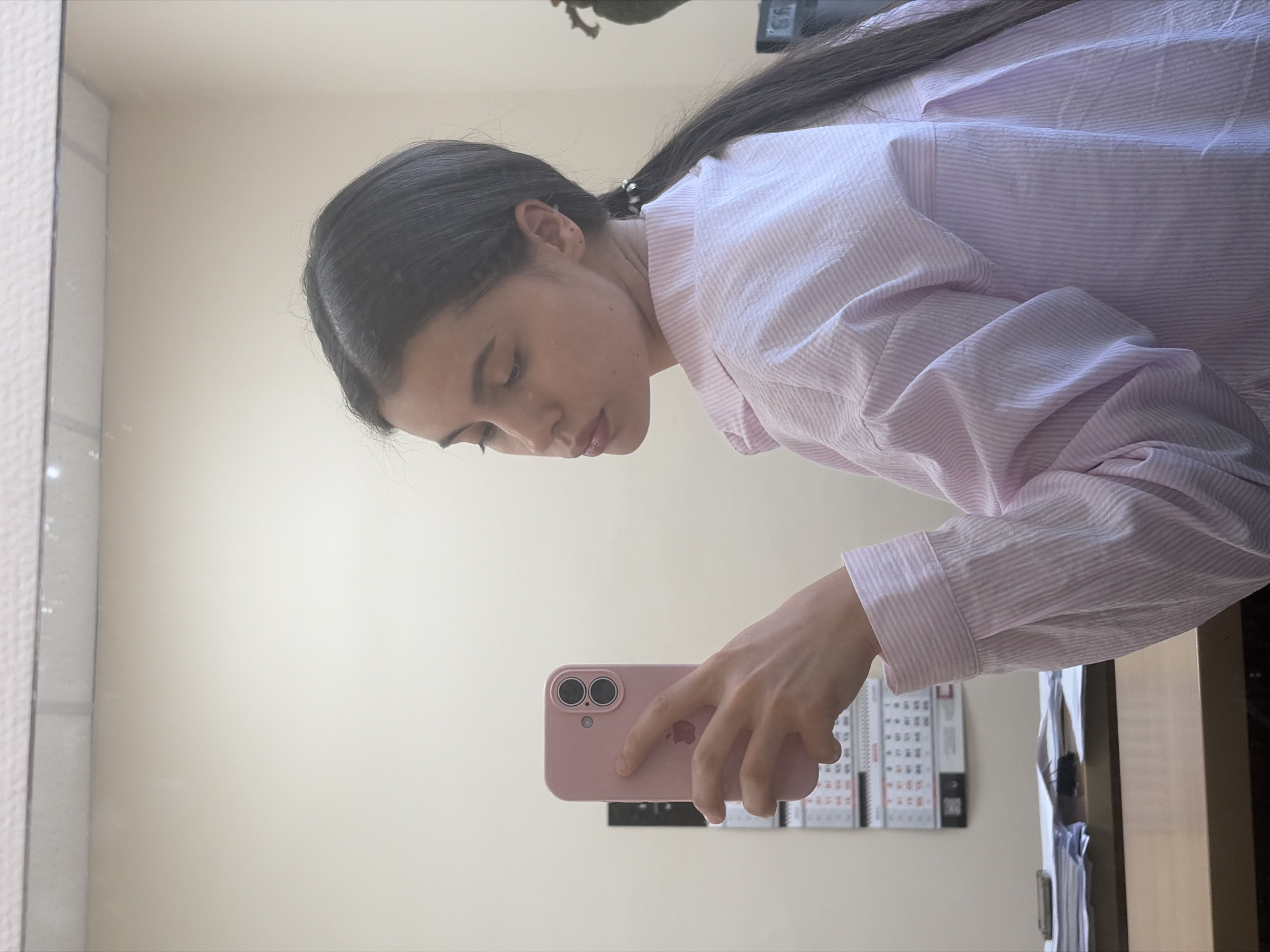 A young woman with long dark hair pulled back into a ponytail is taking a selfie in a mirror. She is wearing a pink and white striped collared shirt. She is holding a pink smartphone in her right hand. In the background, a calendar is hanging on a light-colored wall, and a desk with papers is visible. The image appears to have been taken indoors, possibly in an office or a room in Chaikovsky, Russia, given the calendar and desk setup.