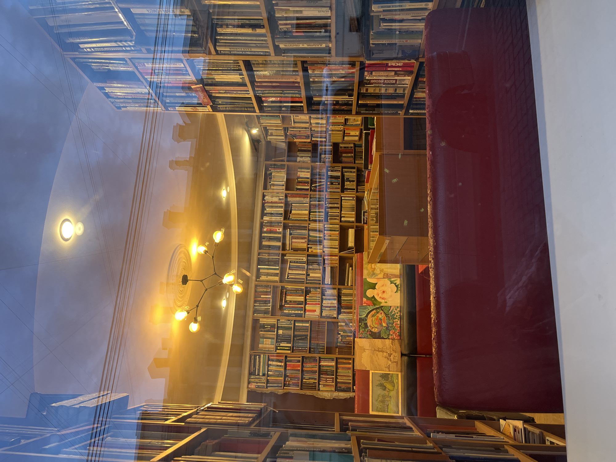 The image appears to be taken through a window, showing the reflection of a ceiling and a chandelier. In the background, there are shelves filled with books. A red cushioned seating area is visible, along with some framed artwork on the shelf. The location is a bookstore.