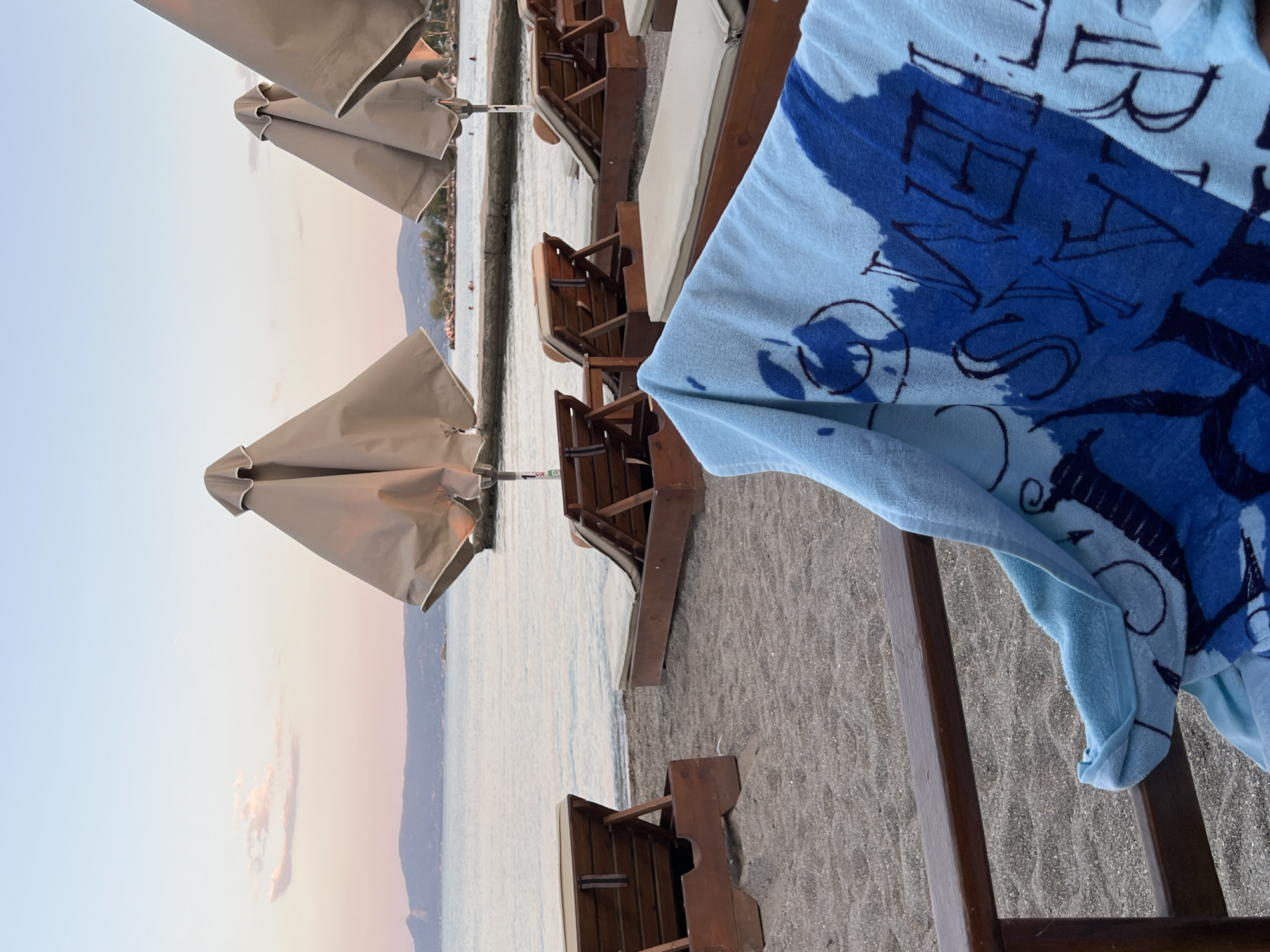 The image depicts a serene beach scene in Amarynthos Municipal Unit, Greece, during what appears to be either sunrise or sunset. In the foreground, a light blue towel with dark blue abstract patterns and lettering is draped over a wooden railing, obscuring part of a wooden chair. Beyond the railing, several empty wooden sun loungers with beige cushions are arranged on a sandy beach, leading towards the calm sea. In the background, closed beach umbrellas stand in a line parallel to the shore. The water is a gentle, reflective expanse, meeting a distant shoreline with hills or mountains. The sky is a soft gradient of pale blue and pink hues, with a few wispy clouds. Several indistinct figures are visible in the shallow water, suggesting the presence of swimmers. The overall atmosphere is peaceful and tranquil.
