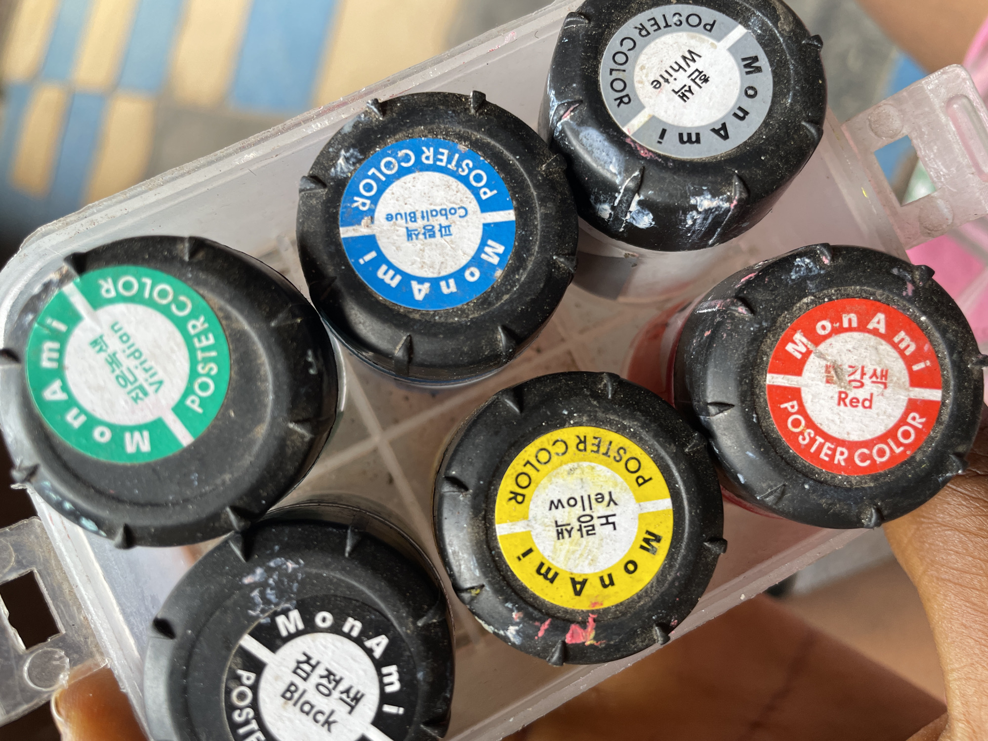 The image shows a close-up view of six small jars of MonAmi poster paint in a clear plastic container. The jars are arranged closely together. Each jar has a black lid with a circular label indicating the color of the paint inside: red, white, yellow, cobalt blue, black, and viridian. The labels feature both English and Korean text, stating the color name. The lids appear slightly worn with remnants of dried paint. A part of the container's handle is visible. The background is blurred and out of focus, but it is partially possible to make out the person holding the container. The location information for Nkpor, Nigeria is not relevant to what is shown in the image, so it is not added to the description.