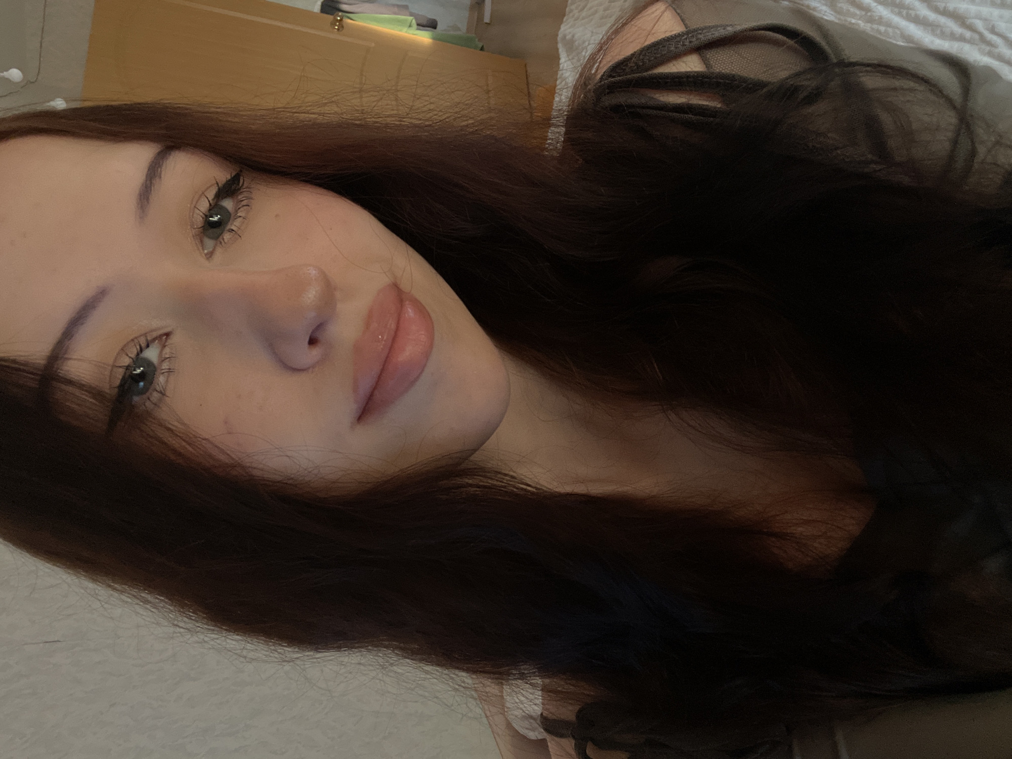 A young female, appearing to be in her late teens or early twenties, is depicted in a close-up portrait, gazing directly at the viewer. She has long, dark brown hair, parted centrally, framing her face and extending over her shoulders. Her light-colored eyes are accentuated with winged eyeliner and mascara, while her dark eyebrows are well-defined. Her fair complexion shows subtle freckles across her nose and cheeks. Her full lips are coated with a light pink or peach-toned gloss and are slightly parted in a soft, subtle smile. She is wearing a dark-colored top, possibly a tank top or camisole, with visible straps and a sheer or mesh panel on her left shoulder (viewer's left).
The background on the left consists of a light-colored wall with a textured surface. To the right, a light brown wooden door is partially visible, with small, round, white decorative string lights hanging above it. Faint glimpses of colorful clothing are visible through the slightly ajar door. The lower right foreground reveals a white textured fabric, consistent with a bedsheet or blanket. The scene is illuminated by warm, artificial light, casting soft shadows. The visual information in the image does not provide any identifying details regarding a specific city or country.